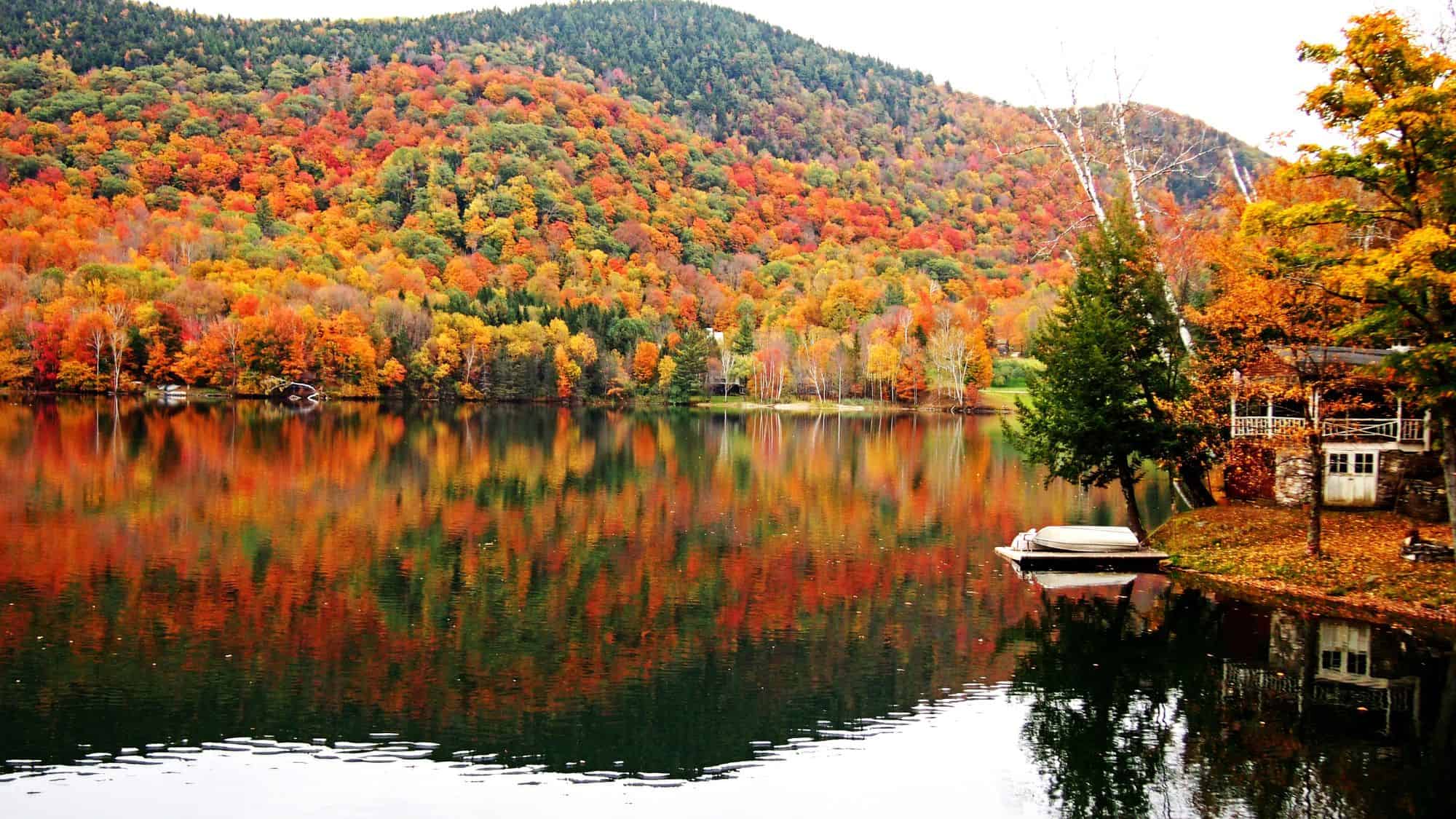 A tranquil lake in Vermont reflecting a forest of vibrant autumn foliage, with shades of red, orange, and yellow, and a small boat docked on the shore.