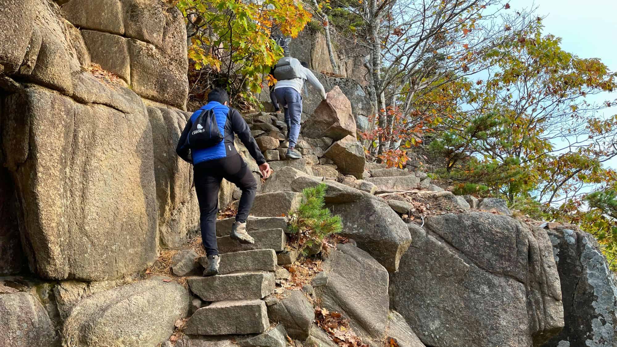 Two hikers ascend a rugged stone staircase along a steep cliffside in Acadia National Park, surrounded by autumn foliage and jagged rock formations.