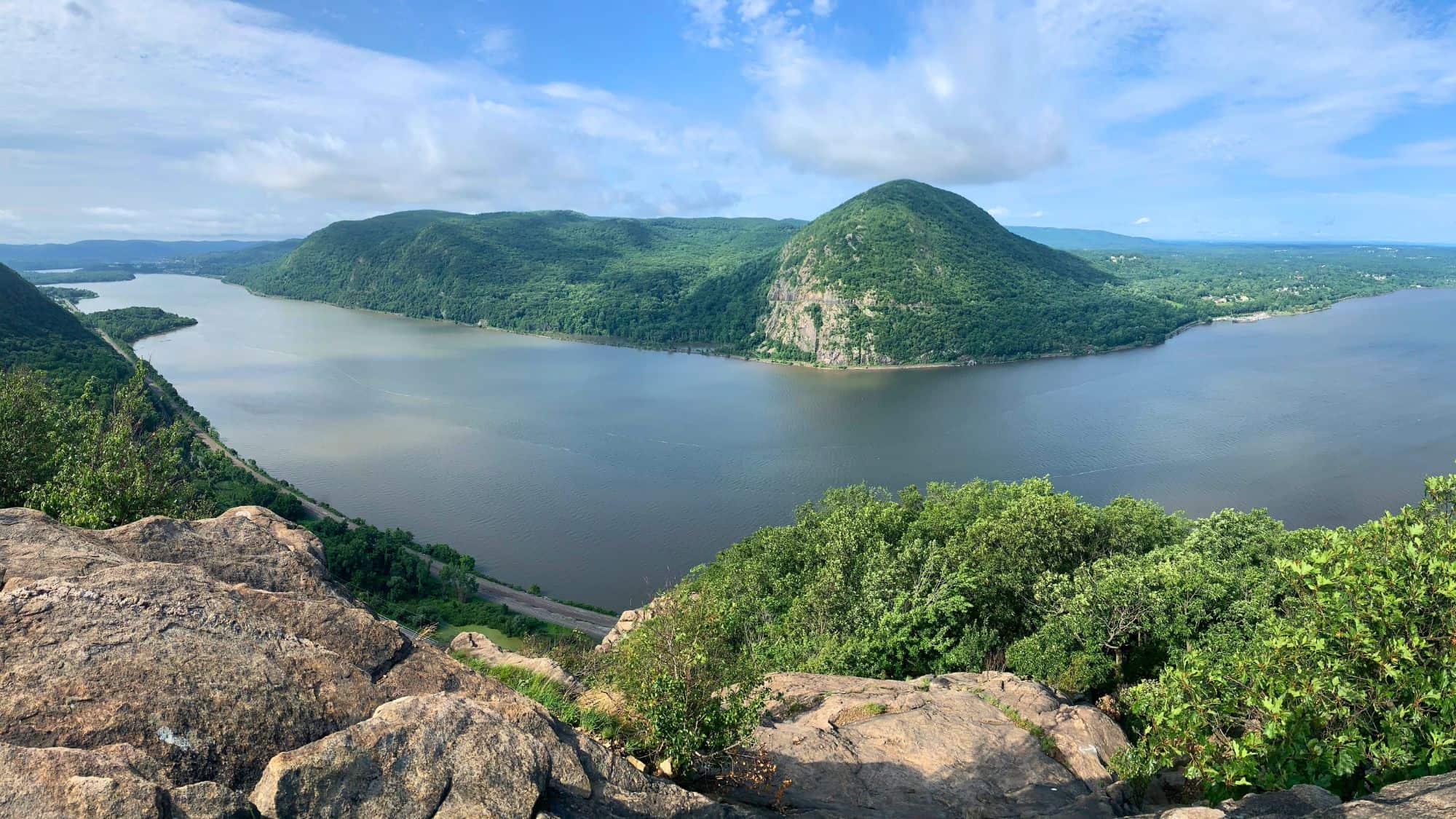 A wide river curves gracefully around a steep, tree-covered hill, viewed from a rocky cliff lined with green shrubs in the foreground. Soft clouds drift across a bright blue sky, highlighting the sweeping, panoramic landscape below.