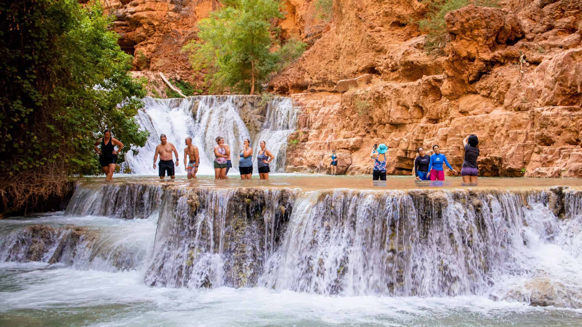 A group of people wade and pose for photos in the terraced cascades of Beaver Falls, with red rock cliffs and lush green trees framing the scene in the Havasupai region of the Grand Canyon.