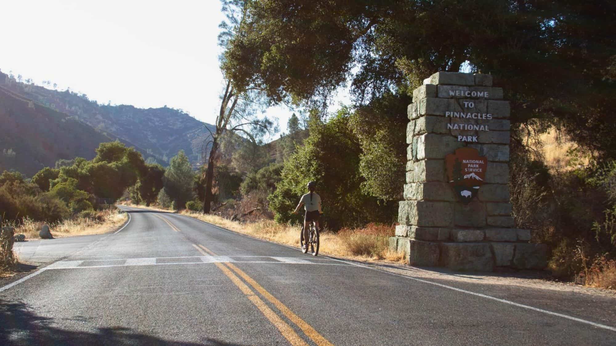 A cyclist rides toward the entrance of Pinnacles National Park, passing a stone sign welcoming visitors, with rolling hills and scattered trees in the background.
