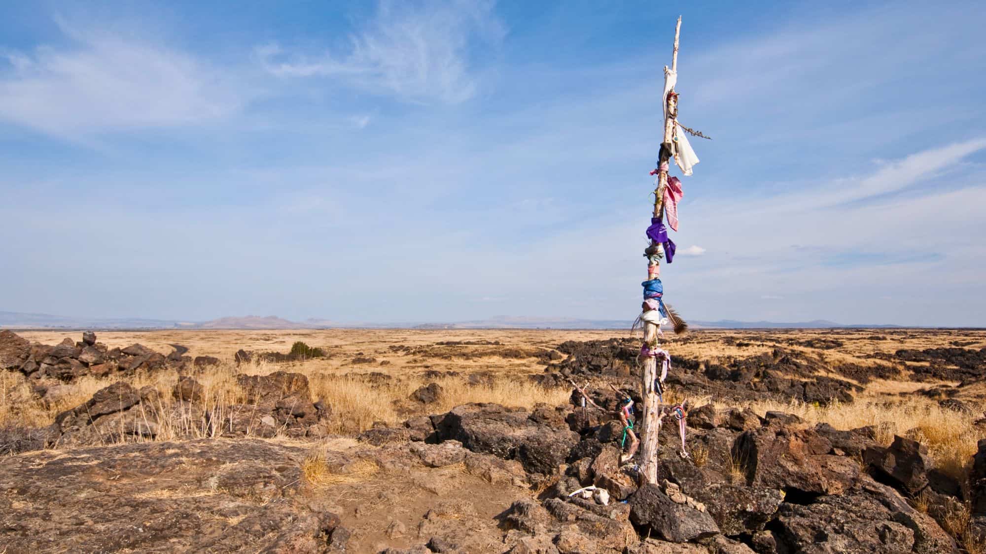 A remote, rugged lava rock landscape with a solitary, decorated wooden pole standing tall in the arid terrain. The pole is adorned with colorful cloth and other offerings, signifying historical or spiritual significance.