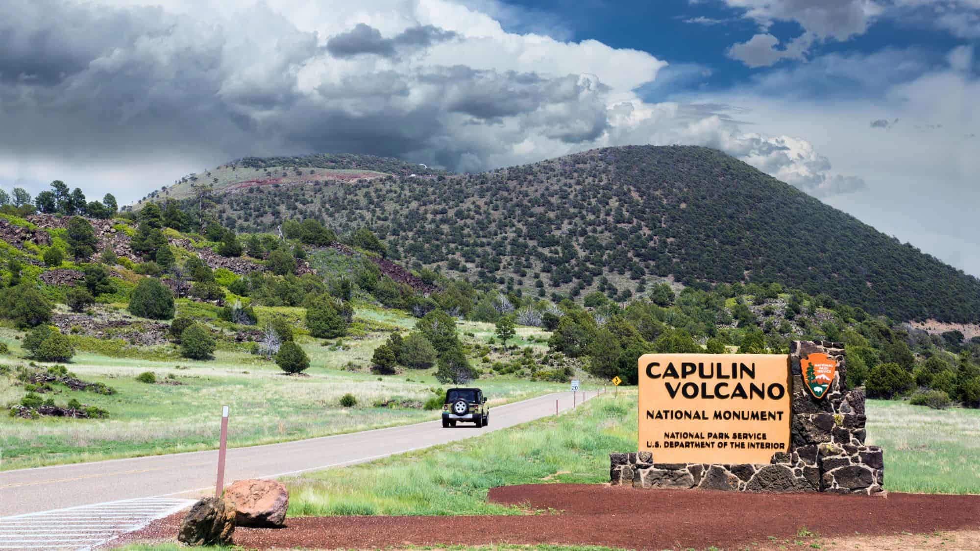 A scenic road leads into Capulin Volcano National Monument, with a green, tree-covered extinct volcanic cone rising in the background and a large stone and metal entrance sign in the foreground under dramatic storm clouds.