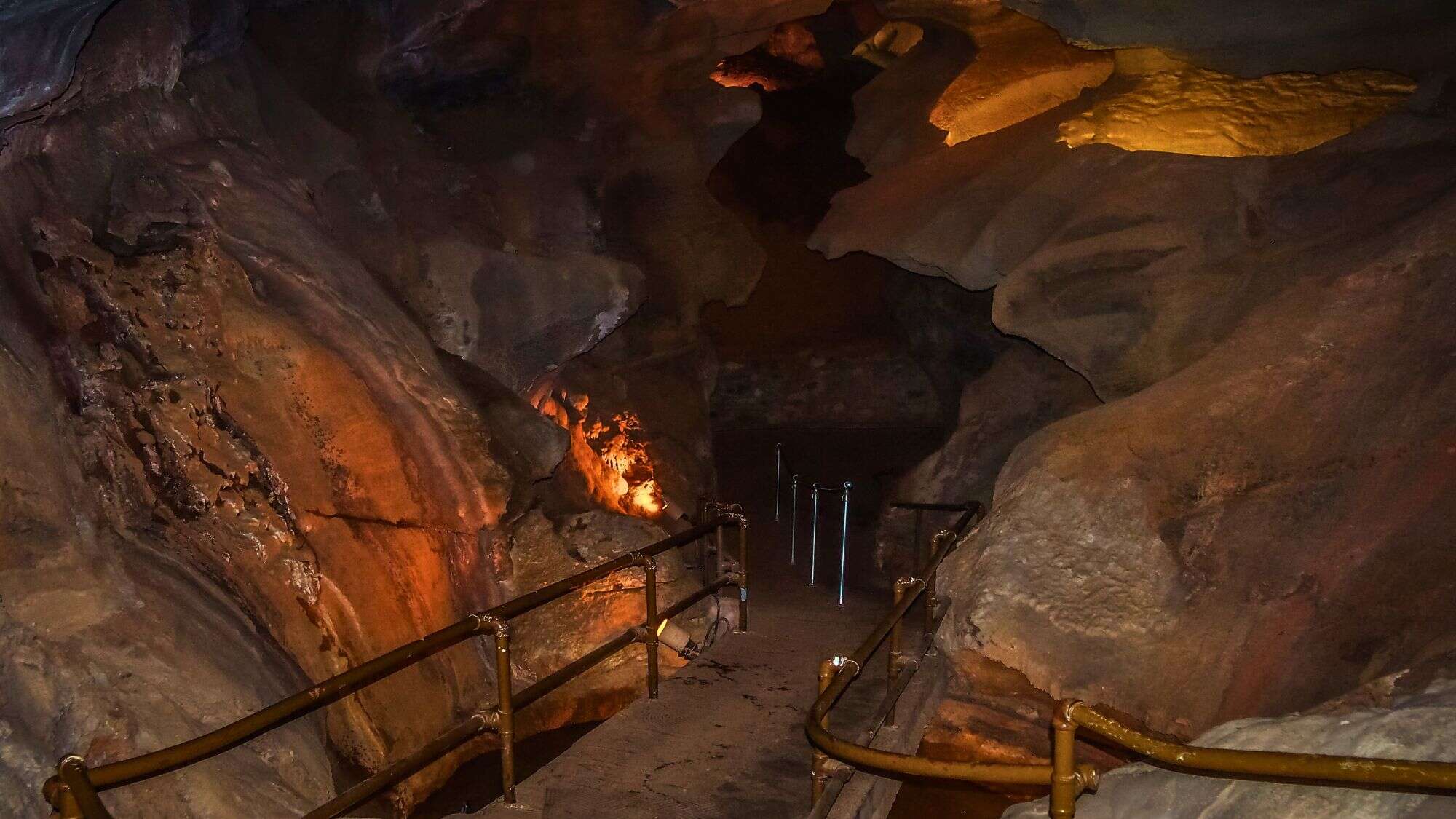 An illuminated view inside Cave of the Winds shows a narrow path with handrails leading through dark, textured rock walls, highlighted by warm amber and gold lighting.