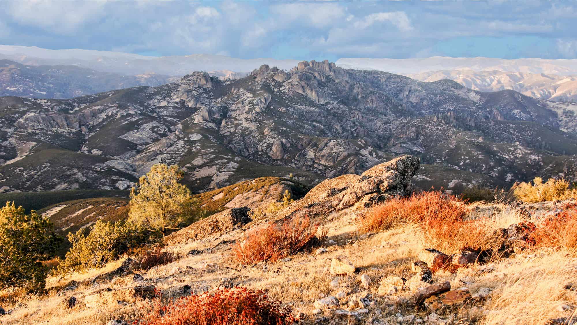 A sweeping view from Chalone Peak, the highest point in Pinnacles National Park, showcasing rugged mountain terrain with dry grass and shrubs bathed in warm sunlight.