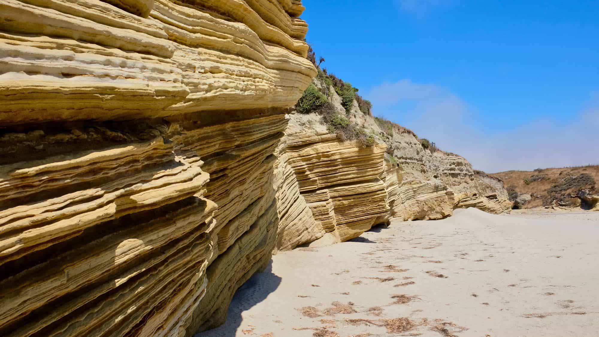 Layered sandstone rock formations rise from a sandy beach, showcasing intricate striations formed over time. The bright blue sky and sparse vegetation contrast against the pale rock.