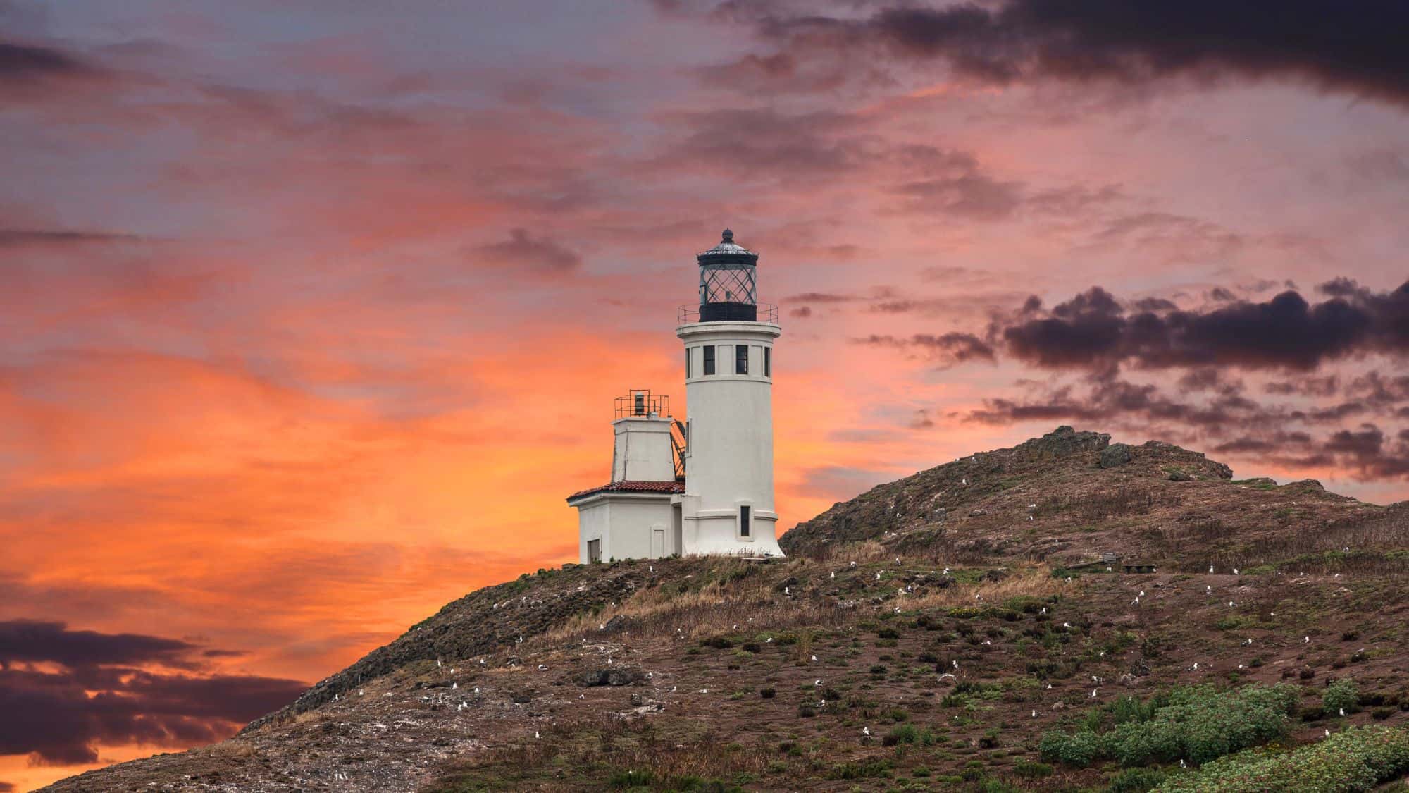 A breathtaking sunset paints the sky in hues of pink, orange, and purple behind a rugged island landscape. A historic lighthouse stands in silhouette against the dramatic backdrop, overlooking the ocean below.