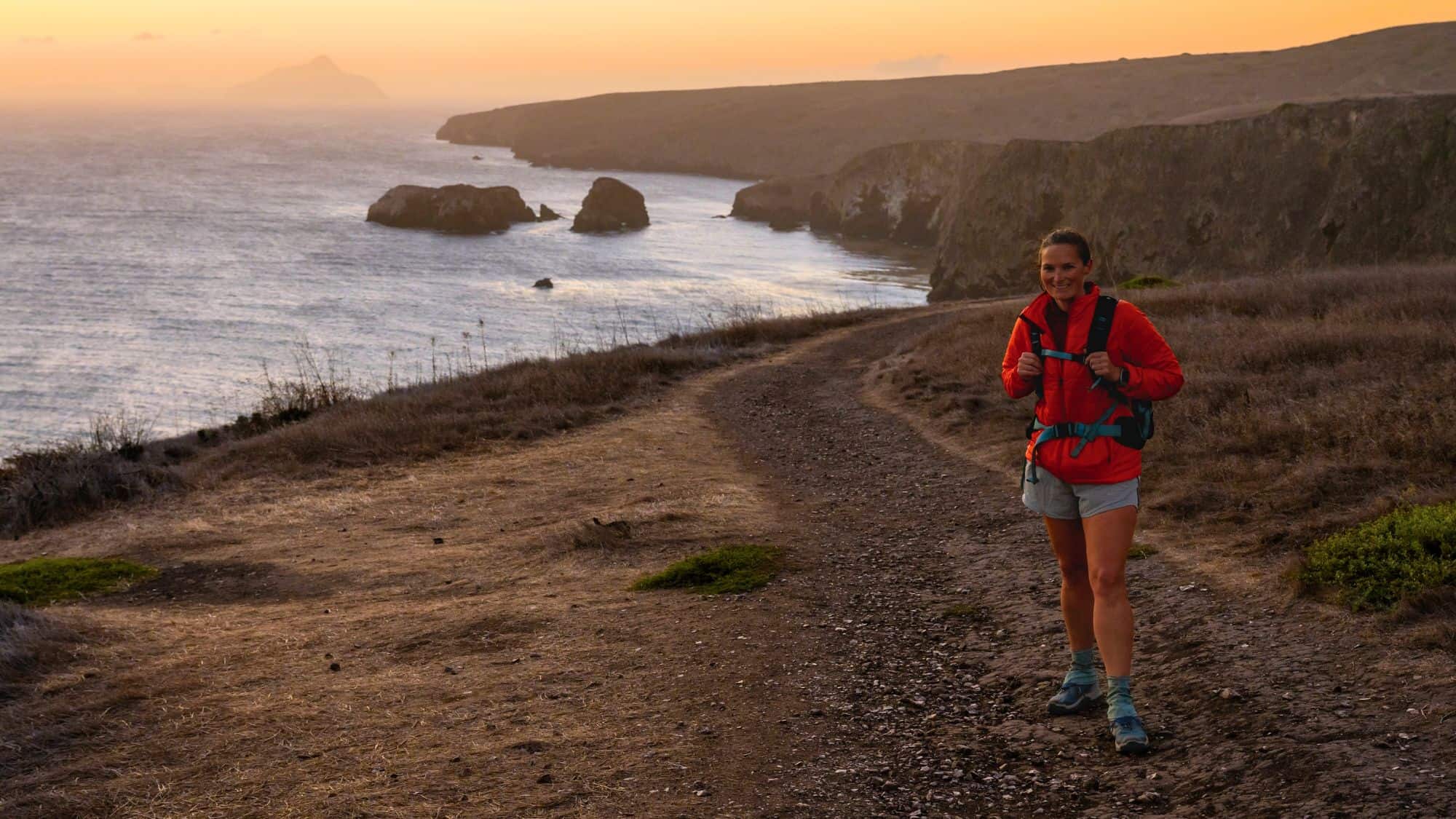 A woman in a red jacket and hiking gear walks along a rugged coastal trail on the Channel Islands at sunset. The cliffs and ocean stretch into the hazy distance, with golden hues reflecting off the water.