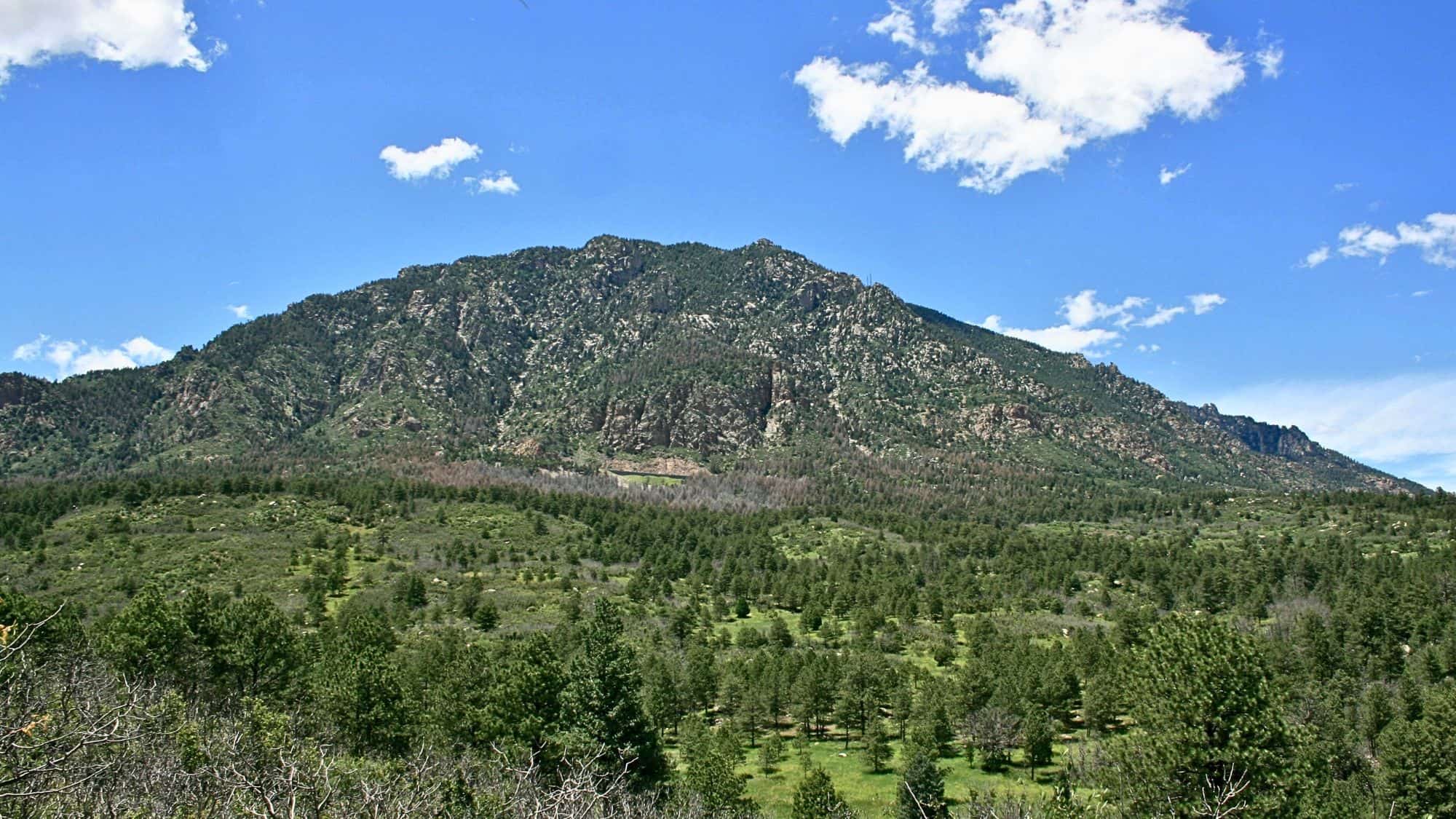 A scenic view of Cheyenne Mountain in Colorado, with its rugged, forest-covered slopes stretching under a clear blue sky dotted with a few fluffy clouds.