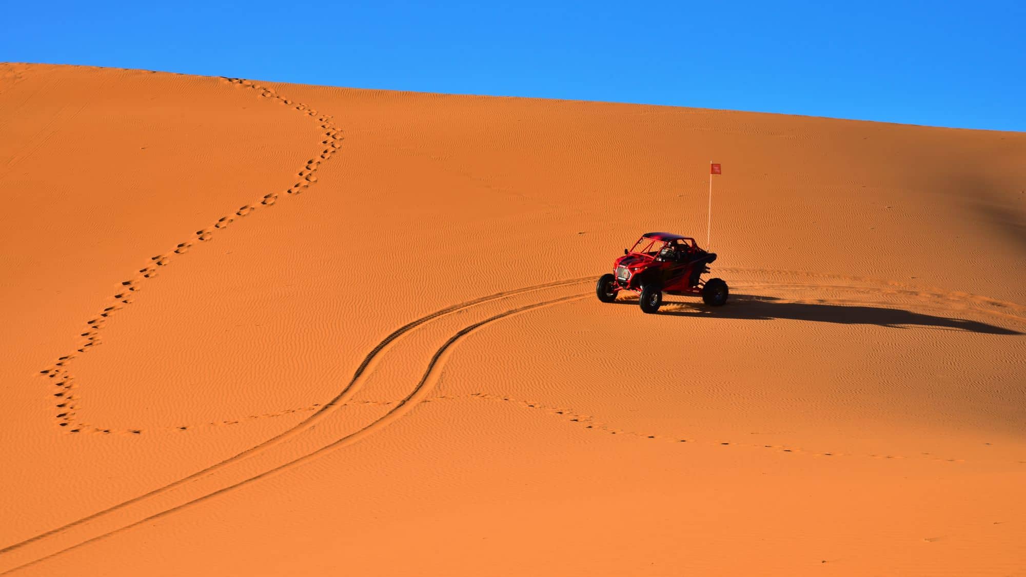 A red all-terrain vehicle (ATV) with a safety flag rides across the vast orange sand dunes, leaving deep tire tracks behind. A set of footprints curves up the slope, contrasting against the untouched sand. The clear blue sky stretches above, adding to the stark beauty of the desert landscape.