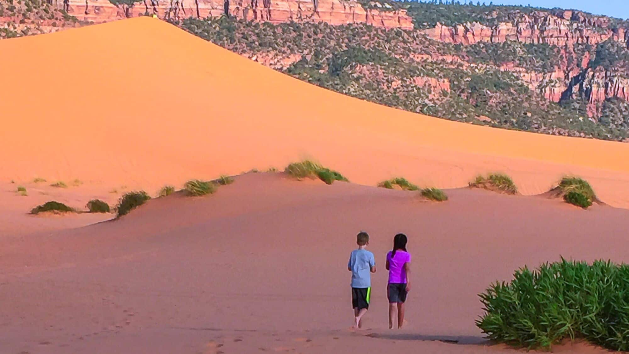 Two children walk barefoot on the warm, golden sand of Coral Pink Sand Dunes, with towering dunes and rugged red cliffs in the background. The late afternoon sun casts soft shadows on the desert floor.