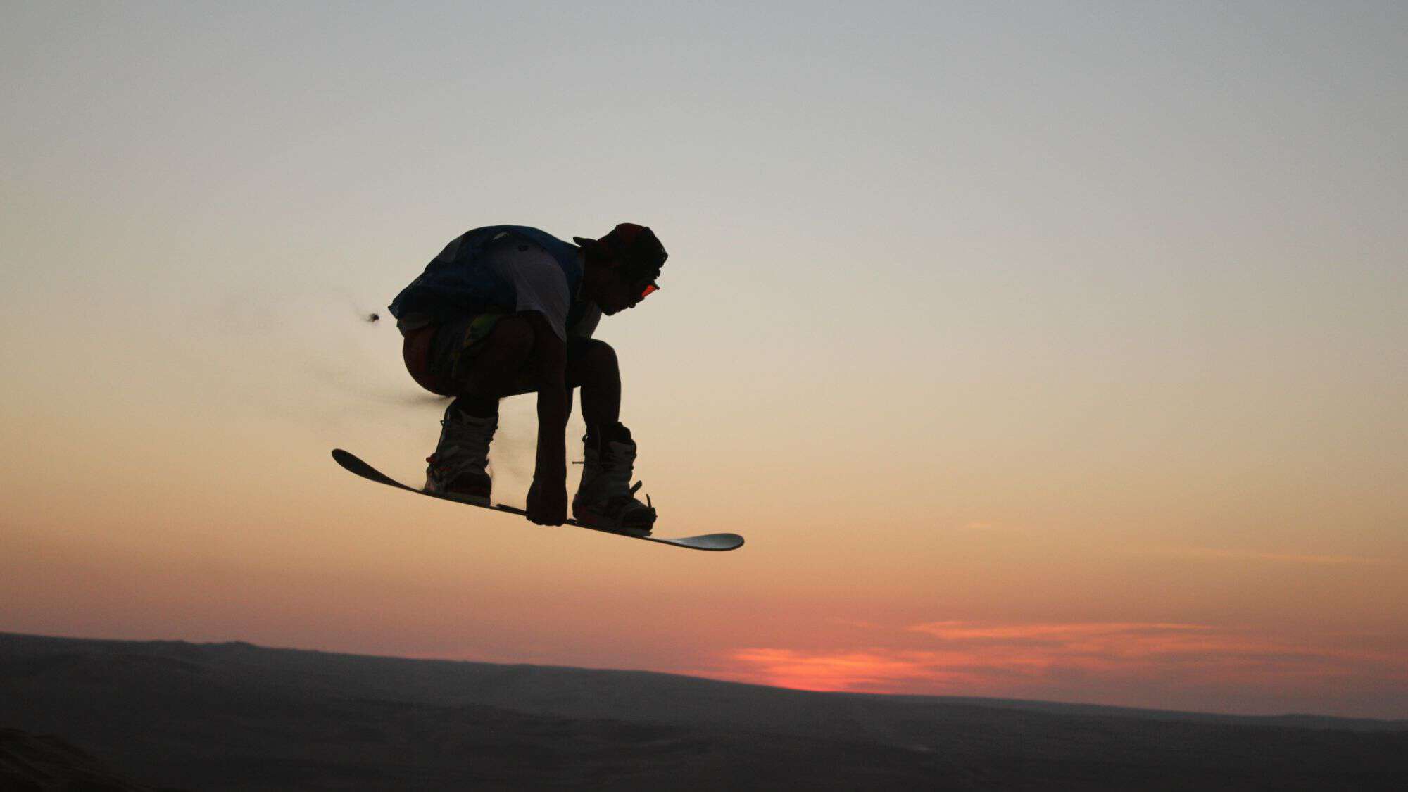 A silhouetted sandboarder catches air against the backdrop of a desert sunset. The rider is mid-jump, with bent knees and arms outstretched, highlighting the thrill of the sport.