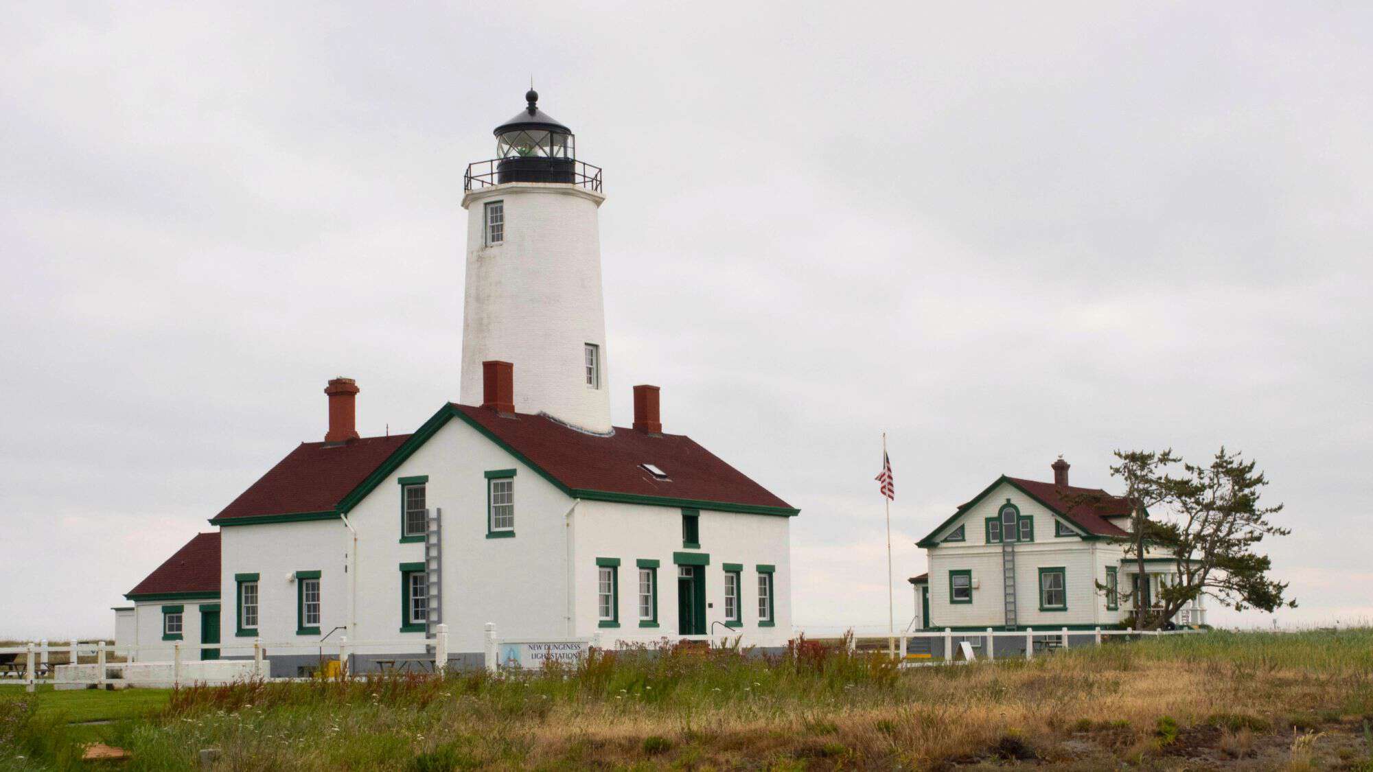 A white lighthouse with a red-roofed keeper's house stands against a cloudy sky. The historic building is surrounded by grassy fields, with a smaller white house and an American flag in the background.