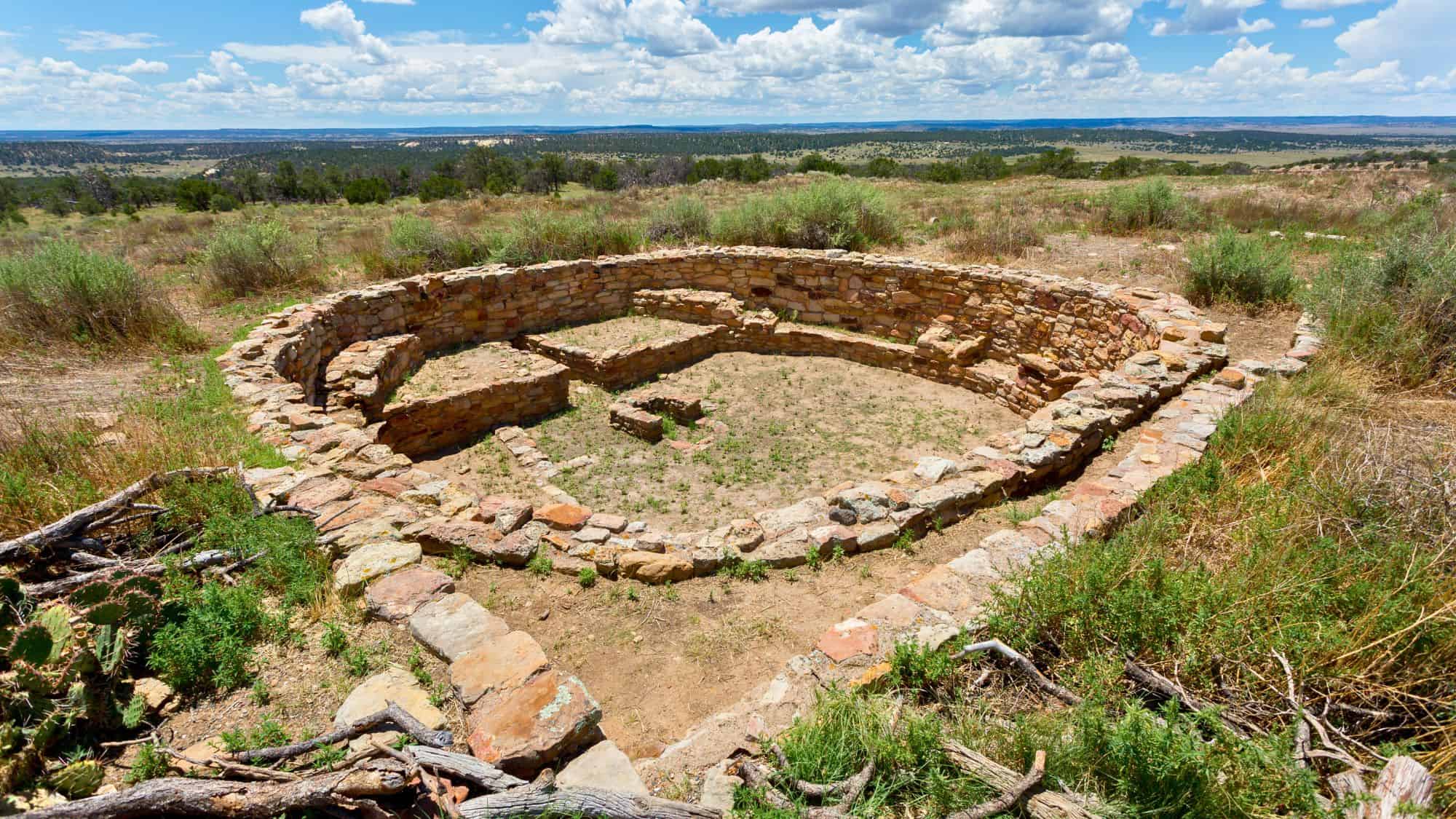 Circular stone ruins from an ancient structure sit among desert vegetation, with wide views of mesas and distant mountains under a sunny sky with scattered clouds.