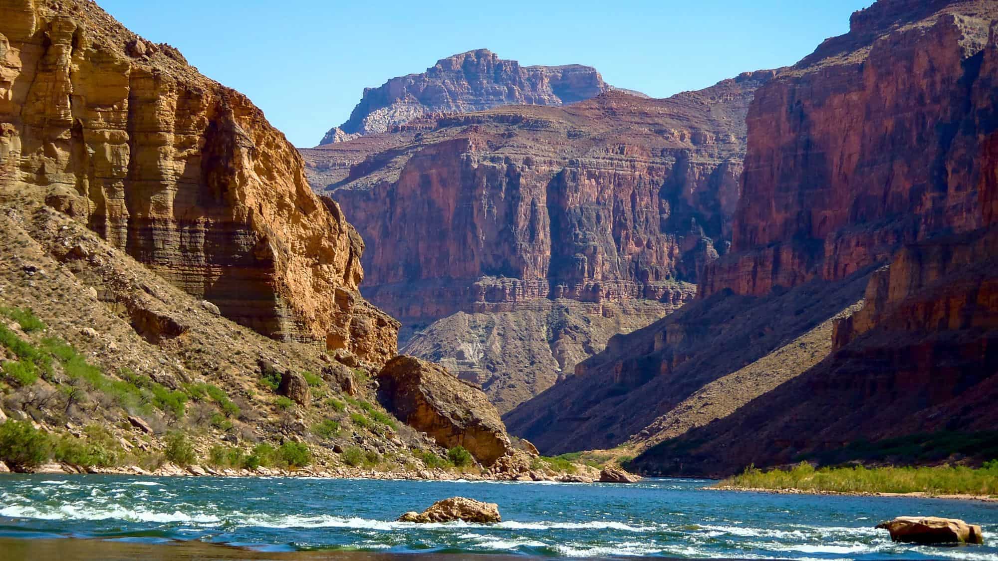 A breathtaking view of the Grand Canyon from the river, with rugged cliffs towering over rushing waters. The reddish rock formations contrast with the bright blue sky.