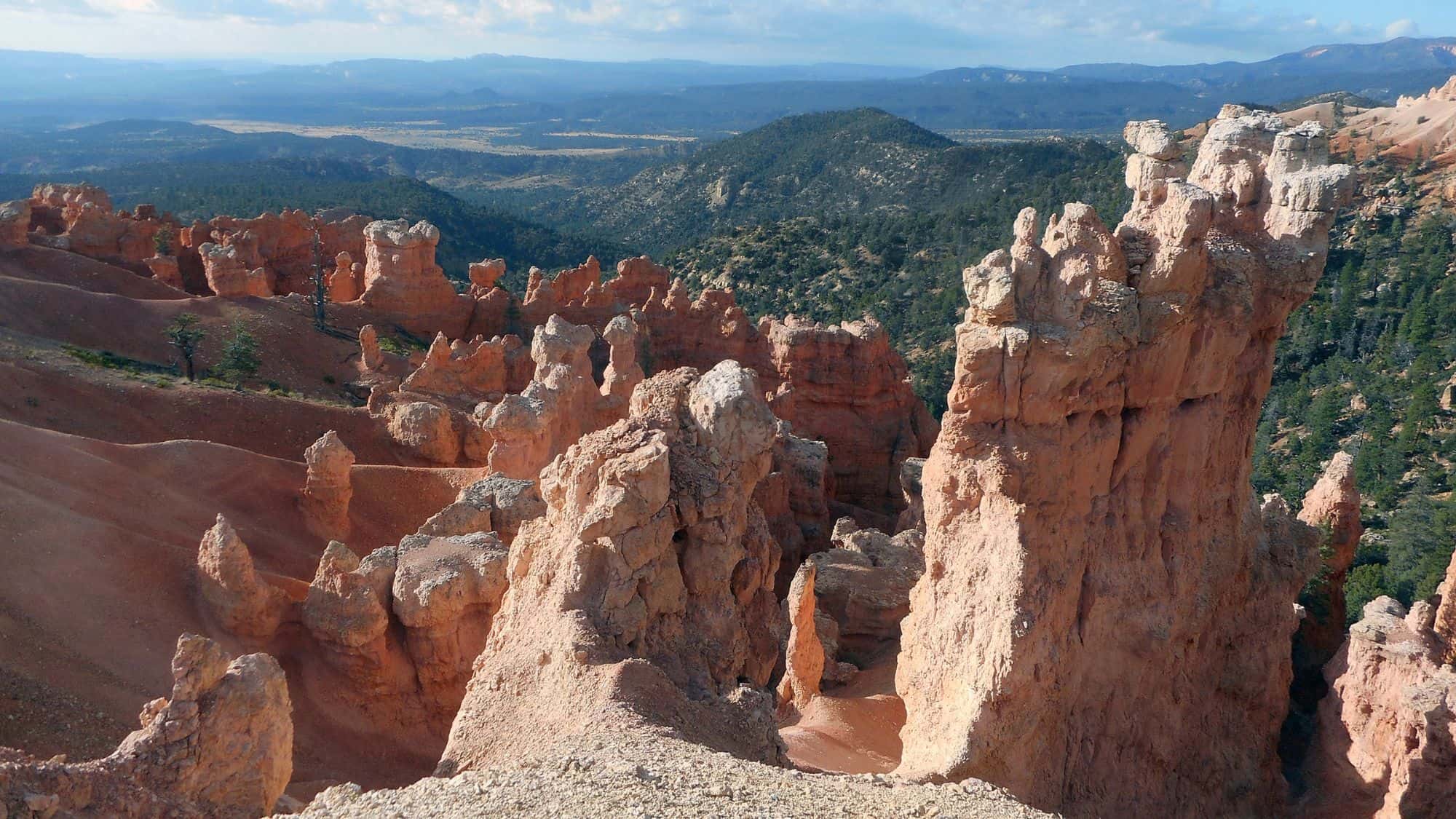 A breathtaking view of Bryce Canyon’s famous rock formations under a clear sky. The towering hoodoos contrast with the deep green forest and rolling hills in the background.