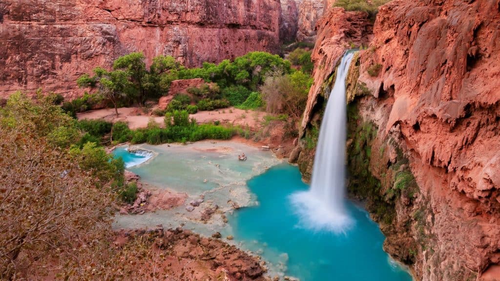 A stunning aerial view of Havasu Falls shows the vibrant turquoise pool and waterfall cascading from red canyon cliffs into a lush desert oasis.