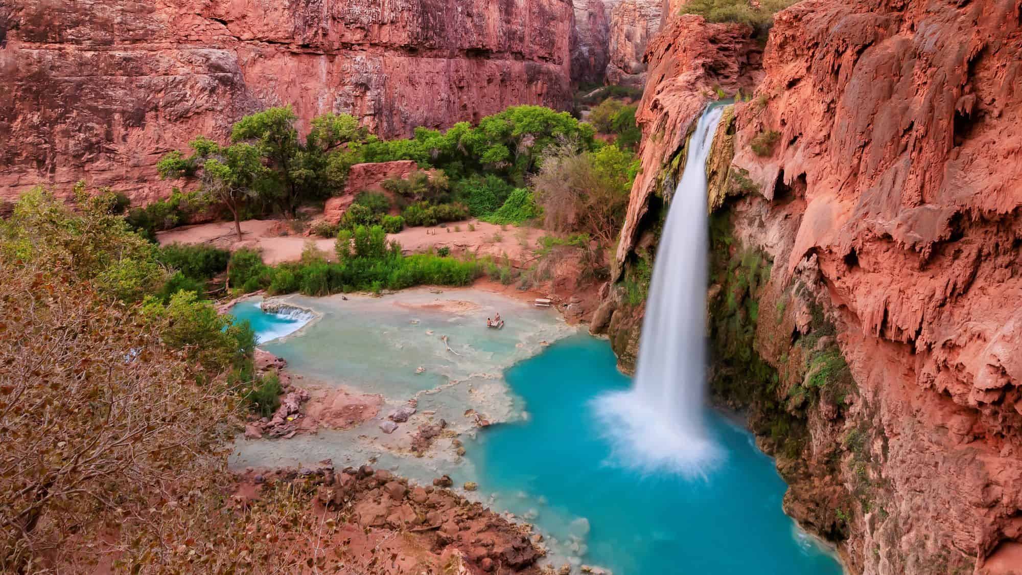 A stunning aerial view of Havasu Falls shows the vibrant turquoise pool and waterfall cascading from red canyon cliffs into a lush desert oasis.