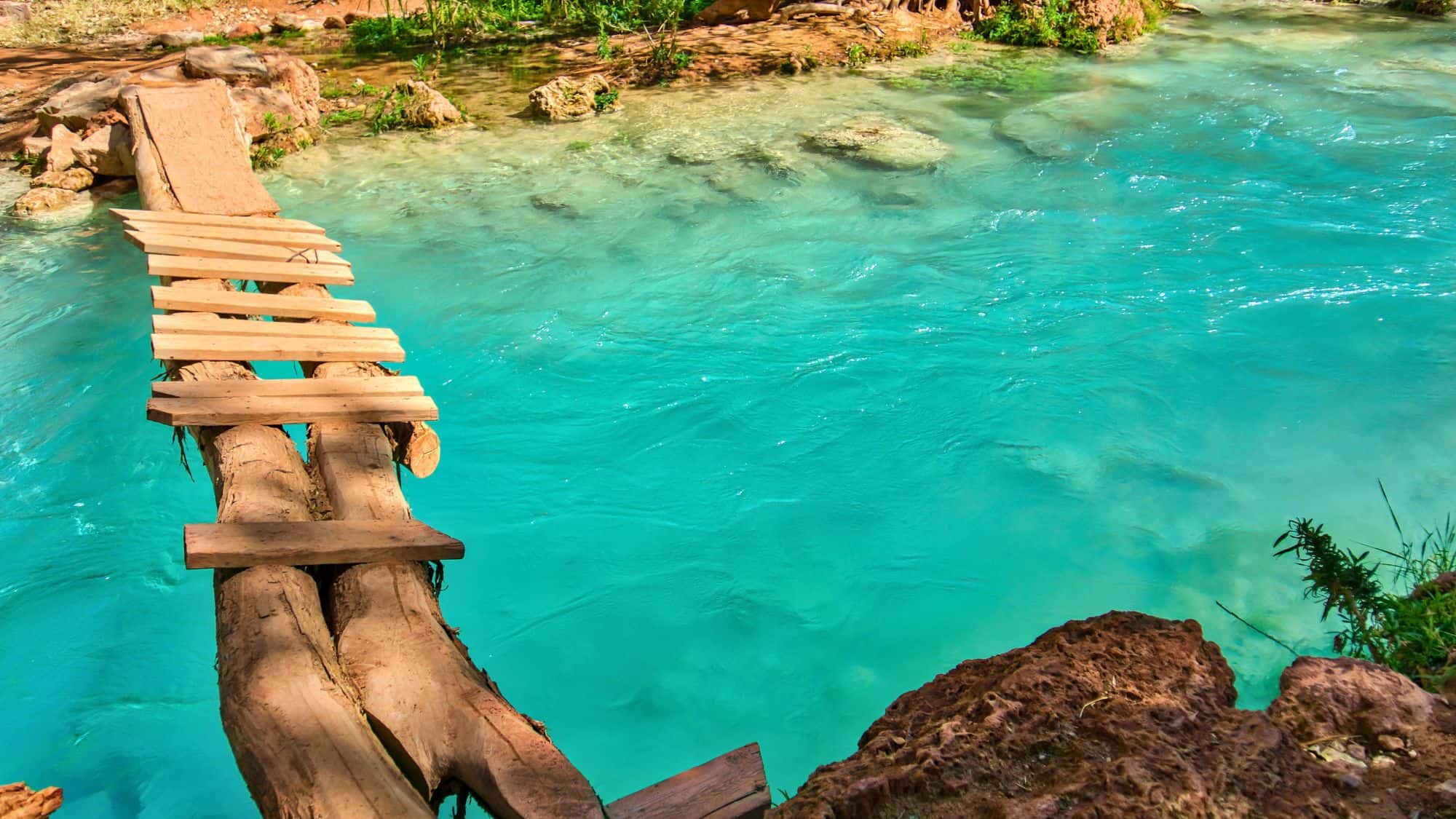 A rustic log and wooden plank bridge stretches over Havasu Creek’s bright turquoise water, set against rocky red terrain and greenery.