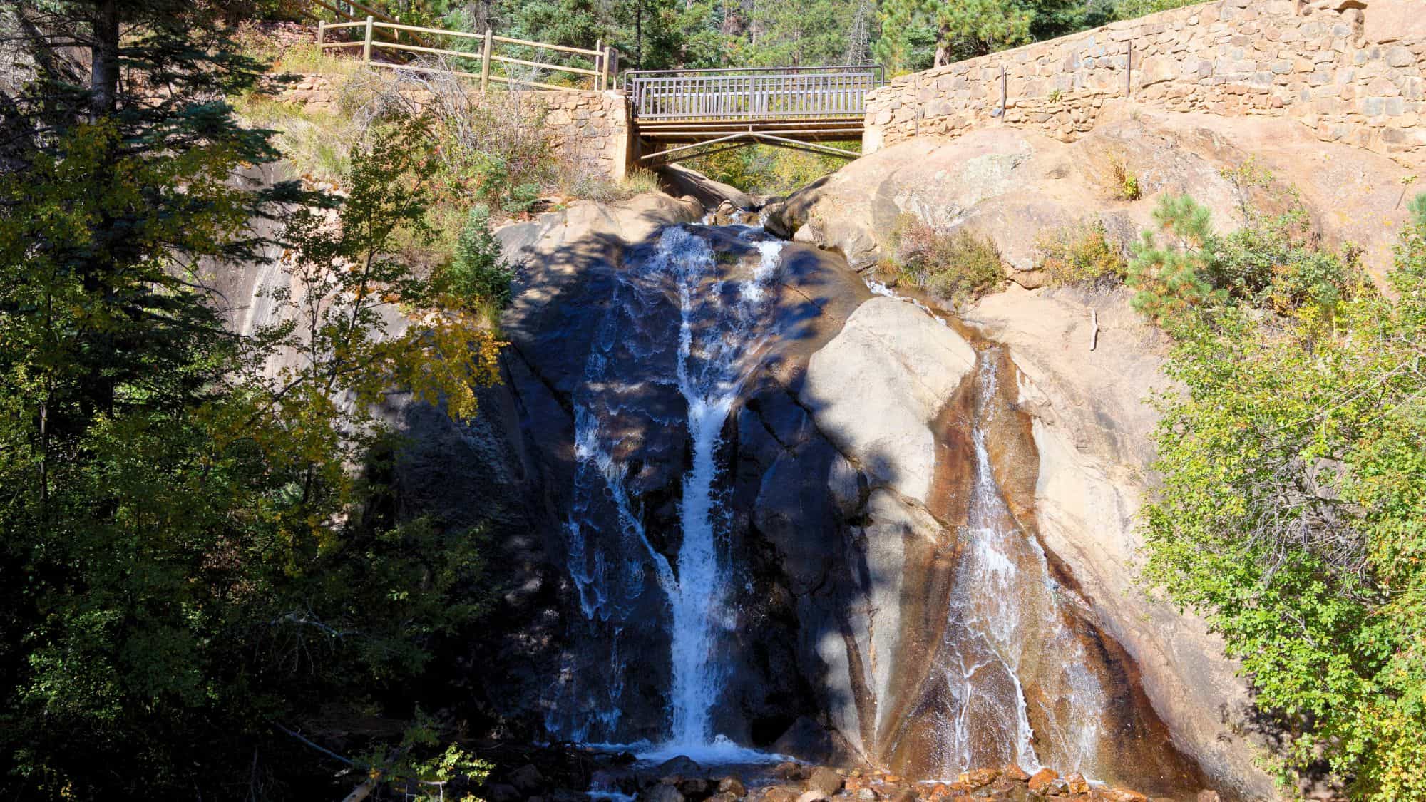 A small but scenic waterfall spills over smooth granite rocks beneath a stone footbridge, surrounded by autumn foliage and evergreens.