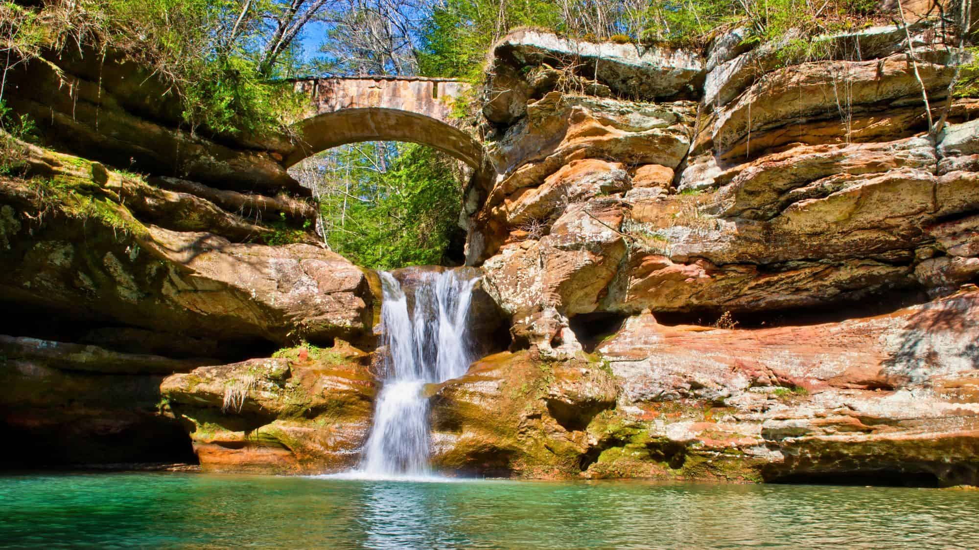 A small but scenic waterfall cascades into a turquoise pool beneath a stone bridge nestled in layered sandstone cliffs, surrounded by green forest in Hocking Hills State Park, Ohio.