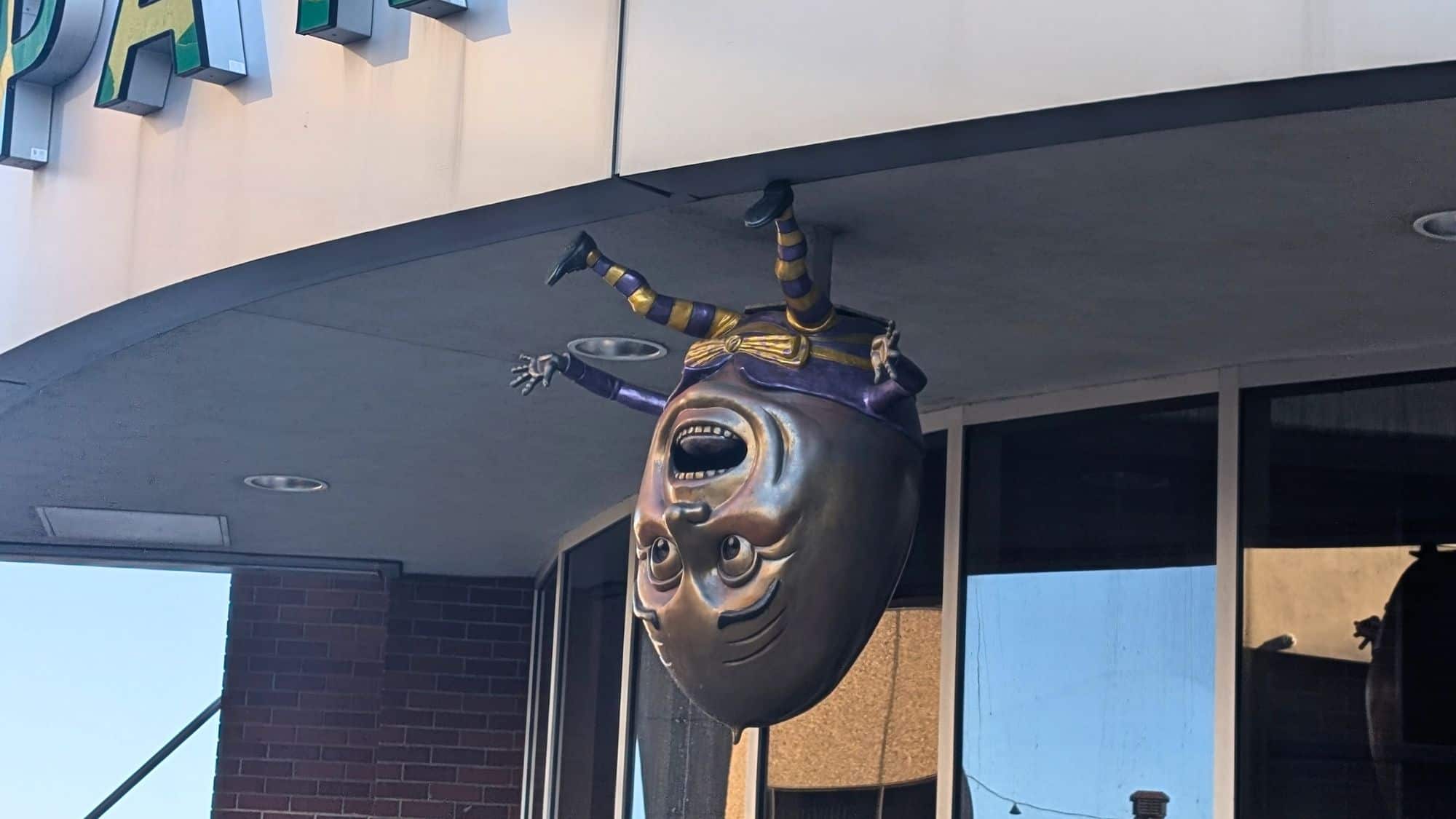 A whimsical sculpture of Humpty Dumpty dressed in purple and yellow, hanging upside down by his feet from a ceiling outside a building, with an exaggerated surprised expression.