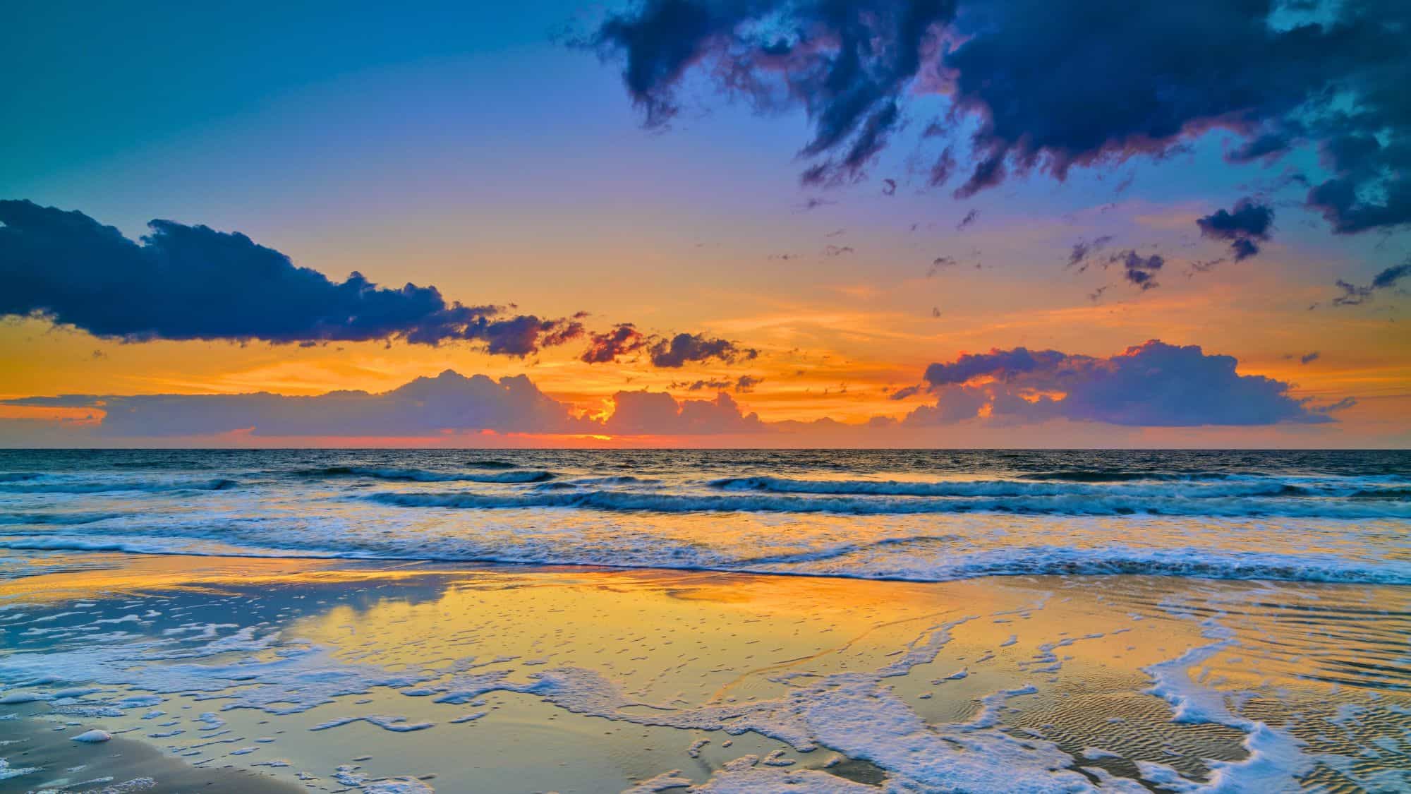 Vivid oranges, purples, and blues light up the sky and ocean at sunrise on the coast of Hunting Island State Park, South Carolina, with soft waves and dramatic clouds completing the peaceful scene.