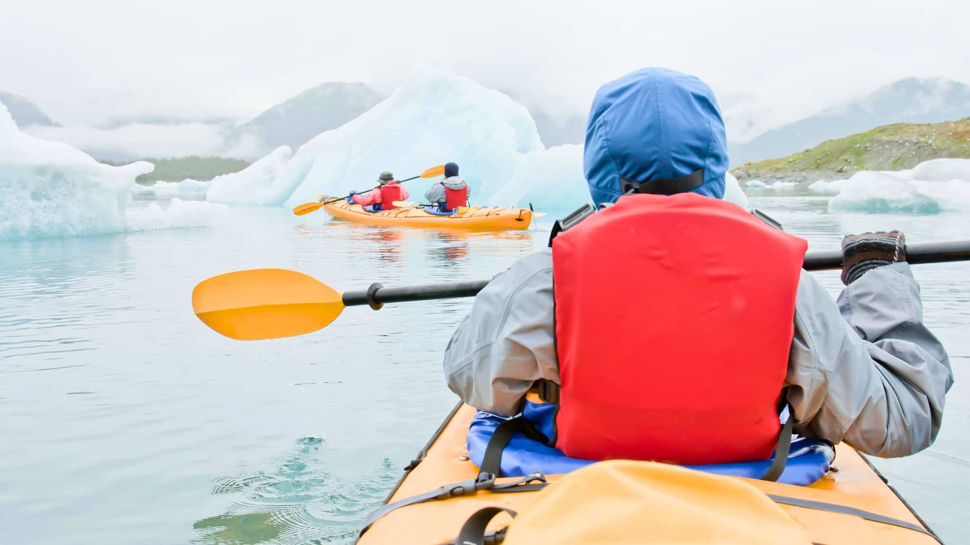 A kayaker in an orange kayak paddles through icy waters surrounded by massive icebergs. The paddler is dressed in cold-weather gear, and misty mountains rise in the distance.