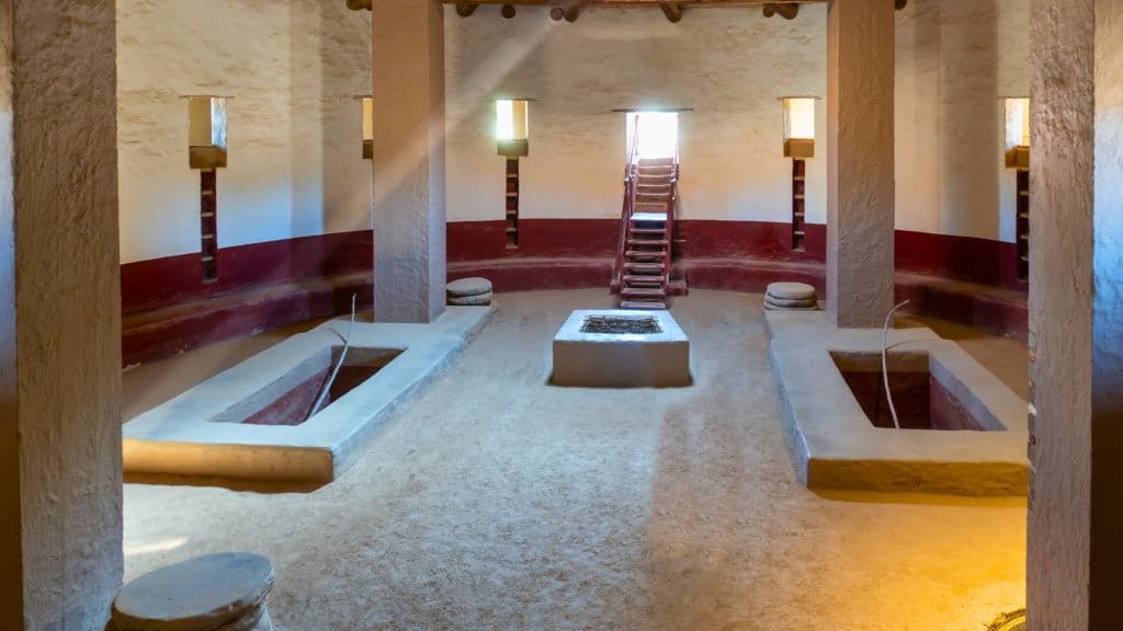 Interior of a reconstructed great kiva, with red and white adobe walls, a central fire pit, roof supports, and benches around the perimeter beneath soft natural lighting.