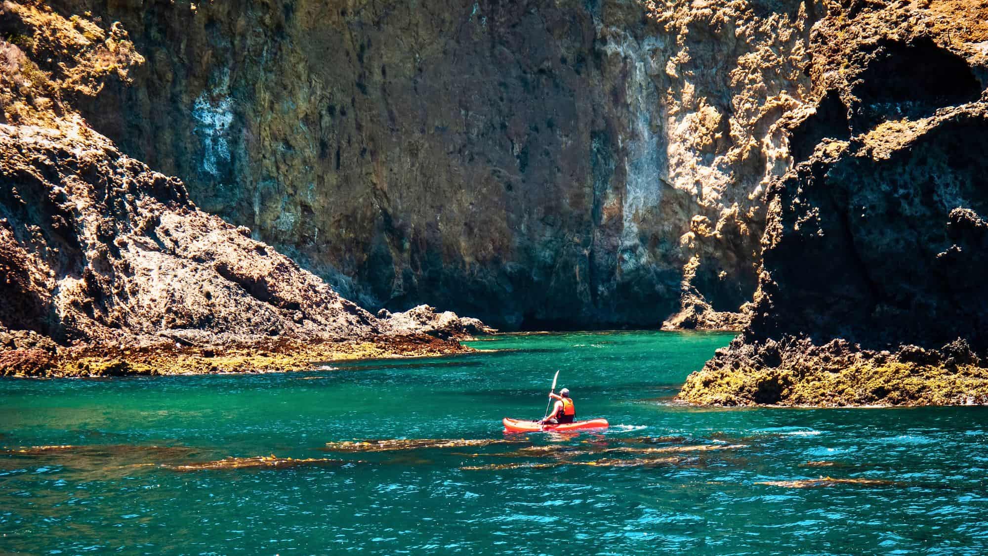 A kayaker in a red life vest paddles through turquoise waters between towering rock cliffs. The sunlight illuminates the rocky formations, creating a dramatic contrast with the water.