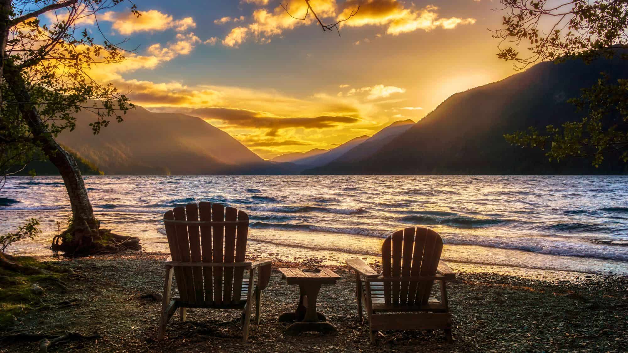 Two wooden Adirondack chairs face Lake Crescent, framed by trees and rippling waters, with mountains catching the last golden light of day.