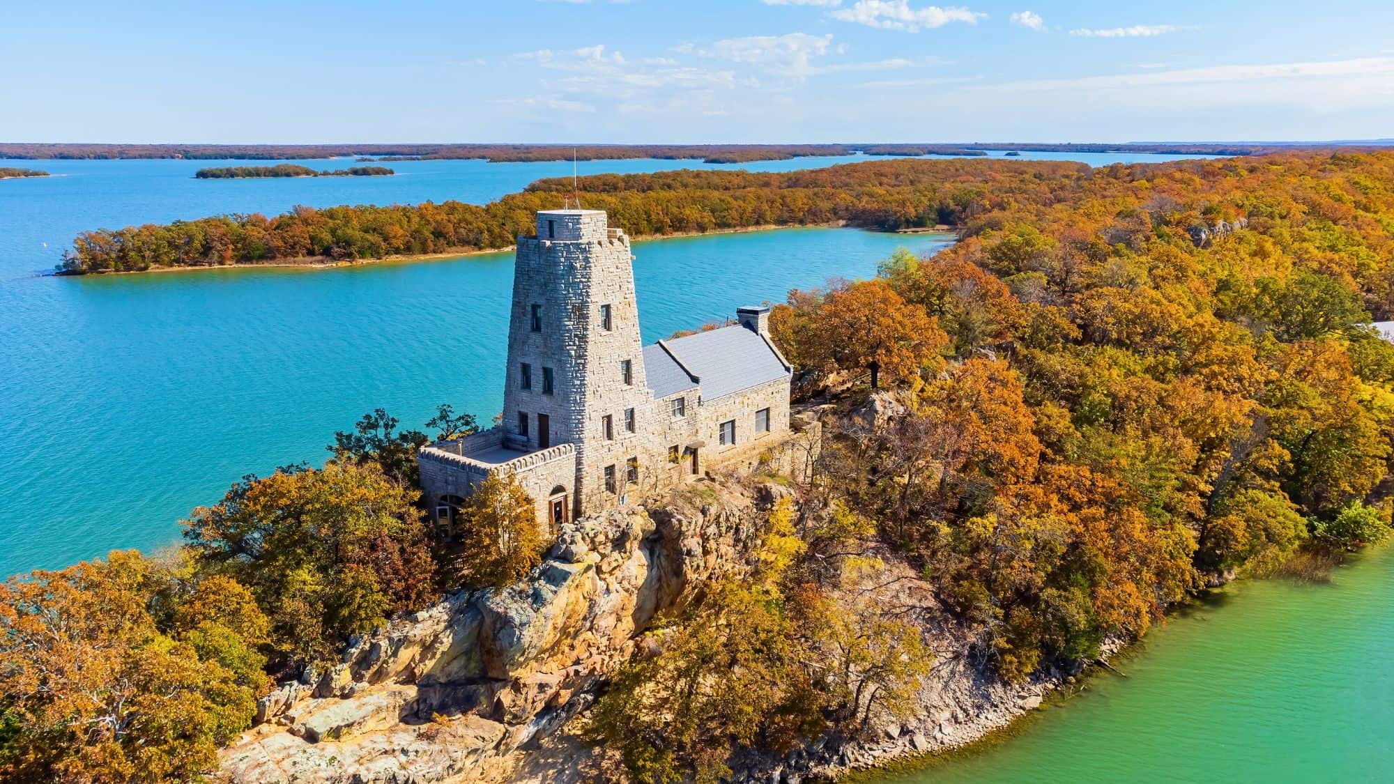 A stone tower-style lodge sits dramatically on a rocky bluff overlooking turquoise waters and colorful fall foliage in Lake Murray State Park, Oklahoma, with tree-covered islands in the distance.