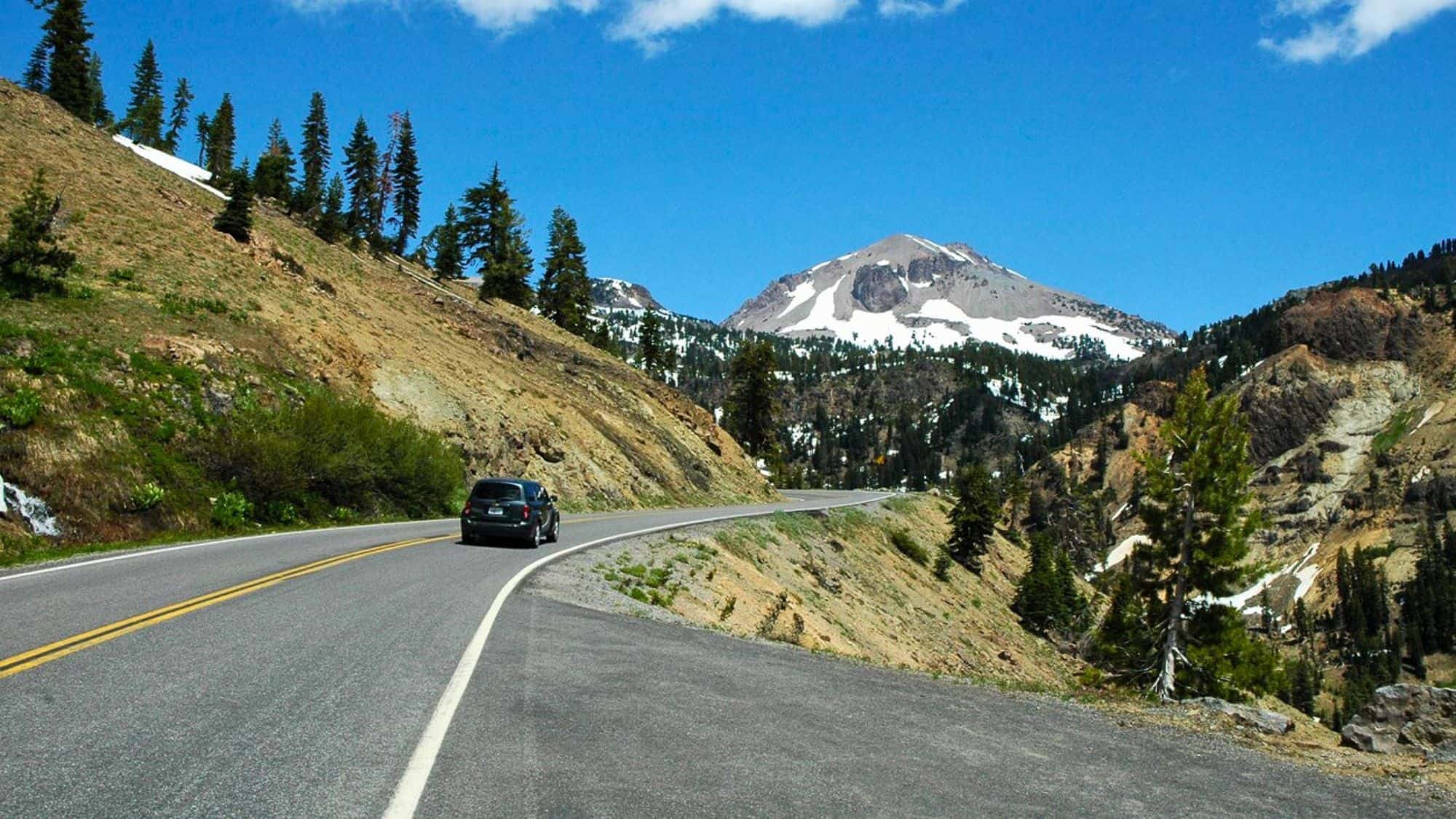 A winding mountain road with a car driving toward a snow-capped peak under a bright blue sky. The landscape features evergreen trees and rugged volcanic terrain.