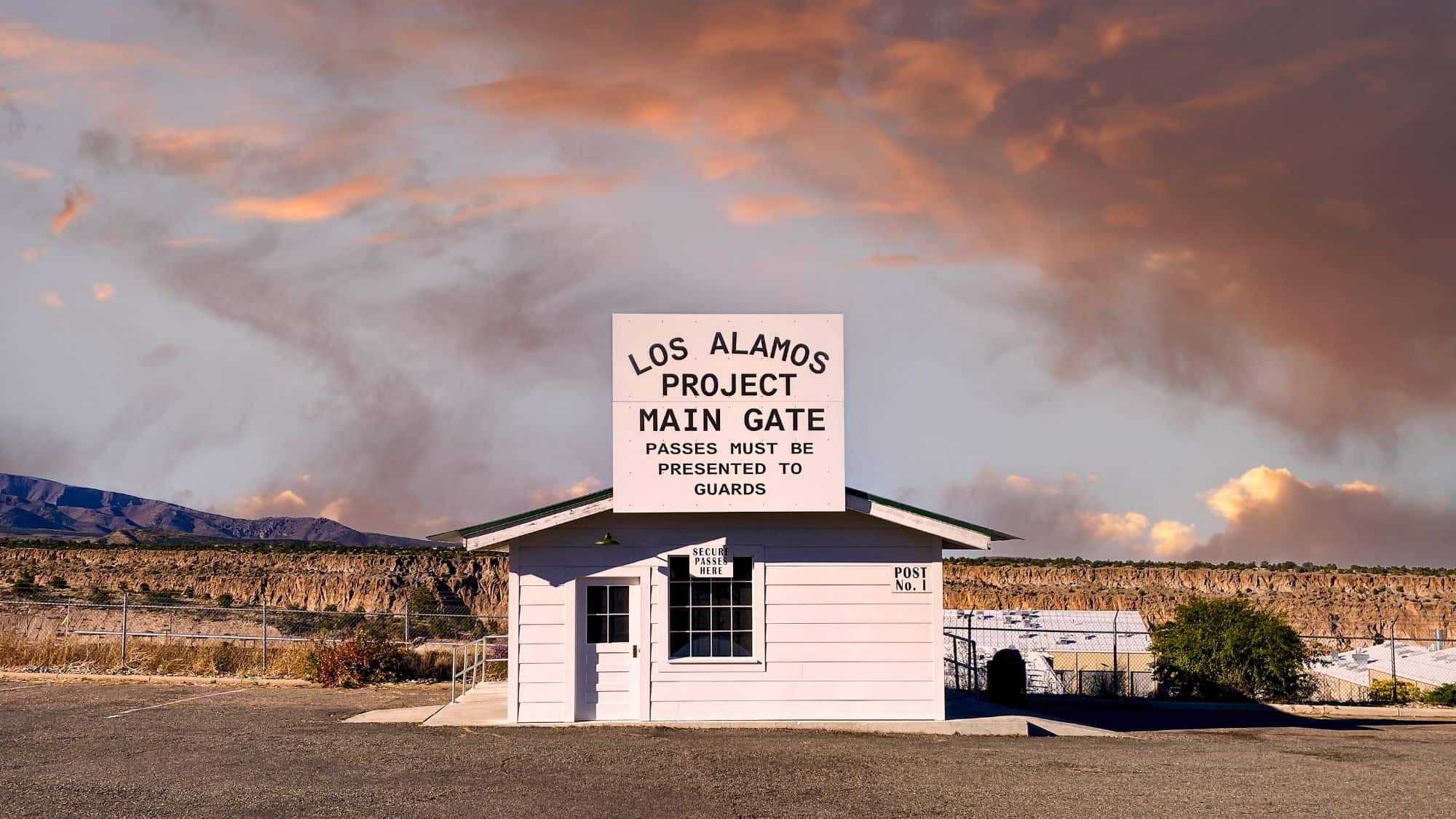 A small white guardhouse marked "Los Alamos Project Main Gate" stands under a dramatic sunset sky, with distant mesas and a chain-link fence surrounding the secured area.