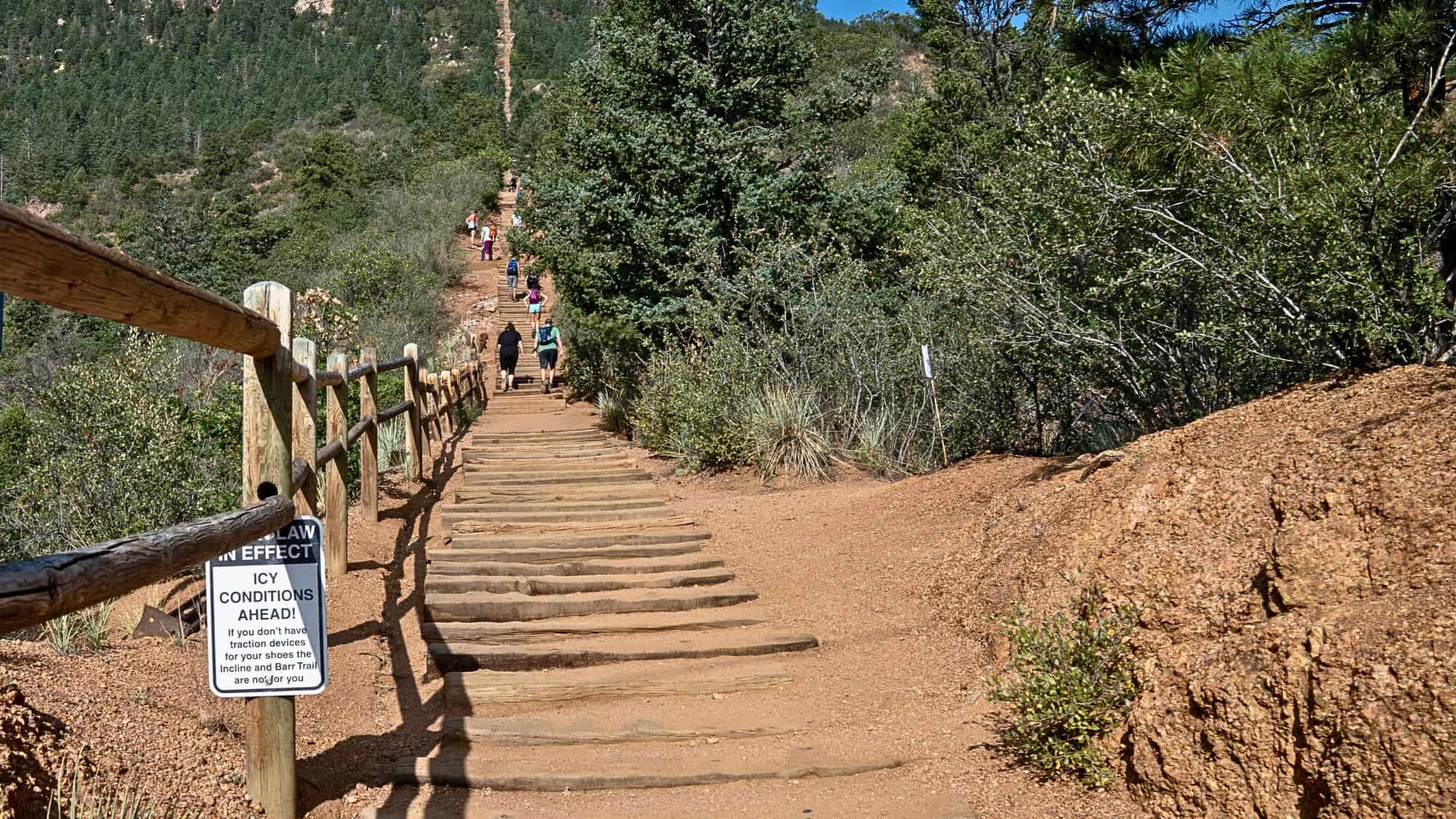 Steep wooden steps form a challenging uphill hiking path flanked by rustic railings and lush greenery, with a warning sign about icy conditions.