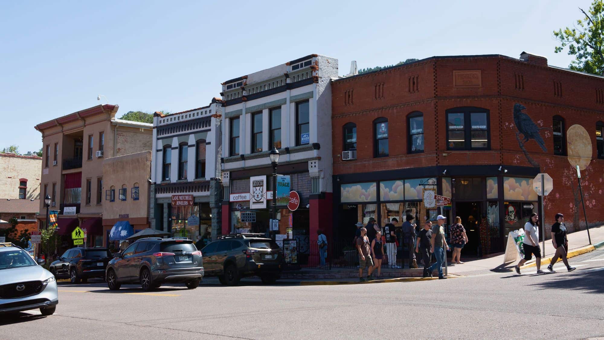 A lively small-town street scene with colorful historic buildings, parked cars, and people strolling past quirky shops and cafés on a sunny day.