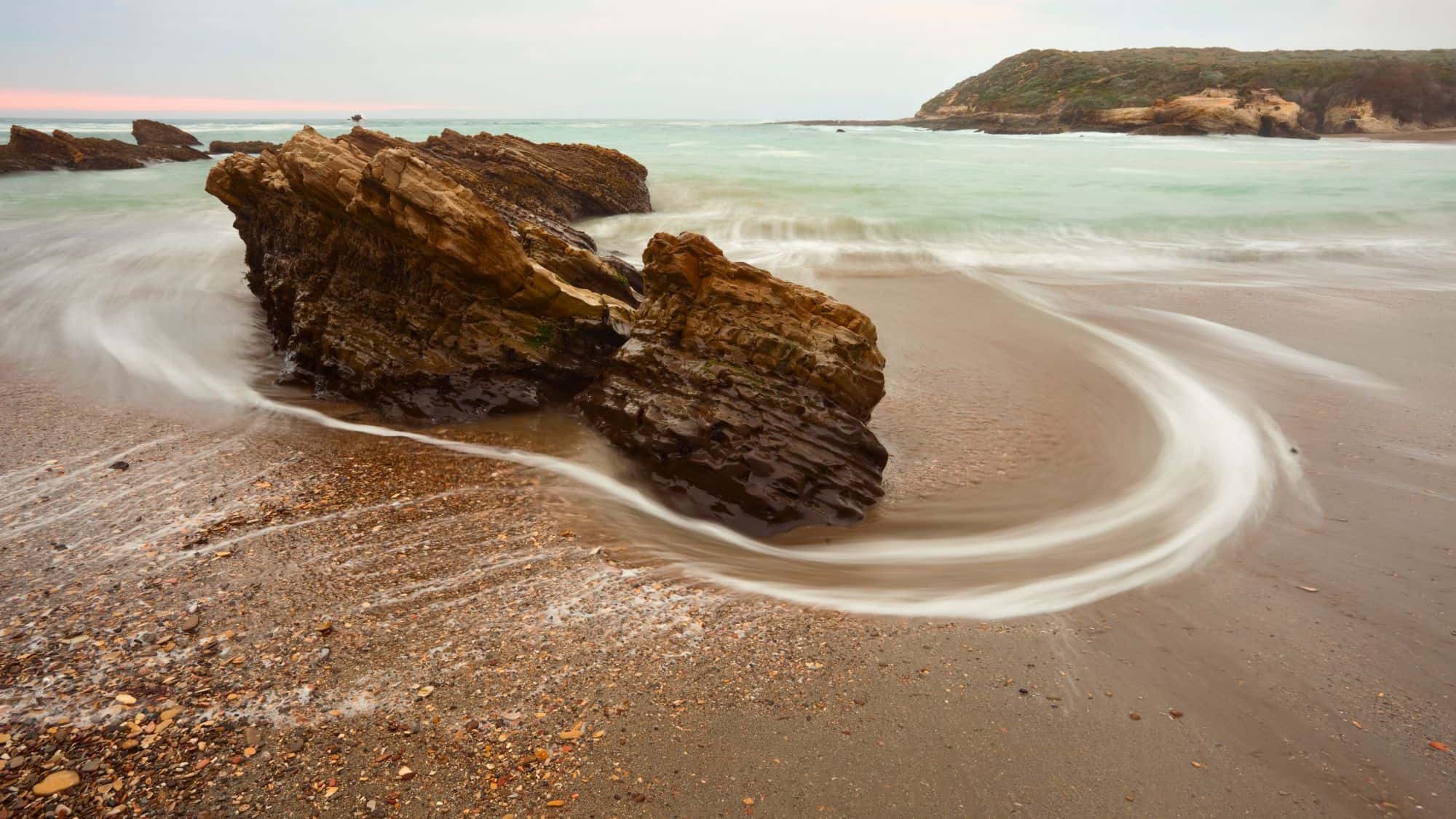 Ocean waves swirl around jagged rock formations on a rugged coastline. The long exposure gives the water a misty, smooth effect as it moves around the rocks, with cliffs and hills in the background.
