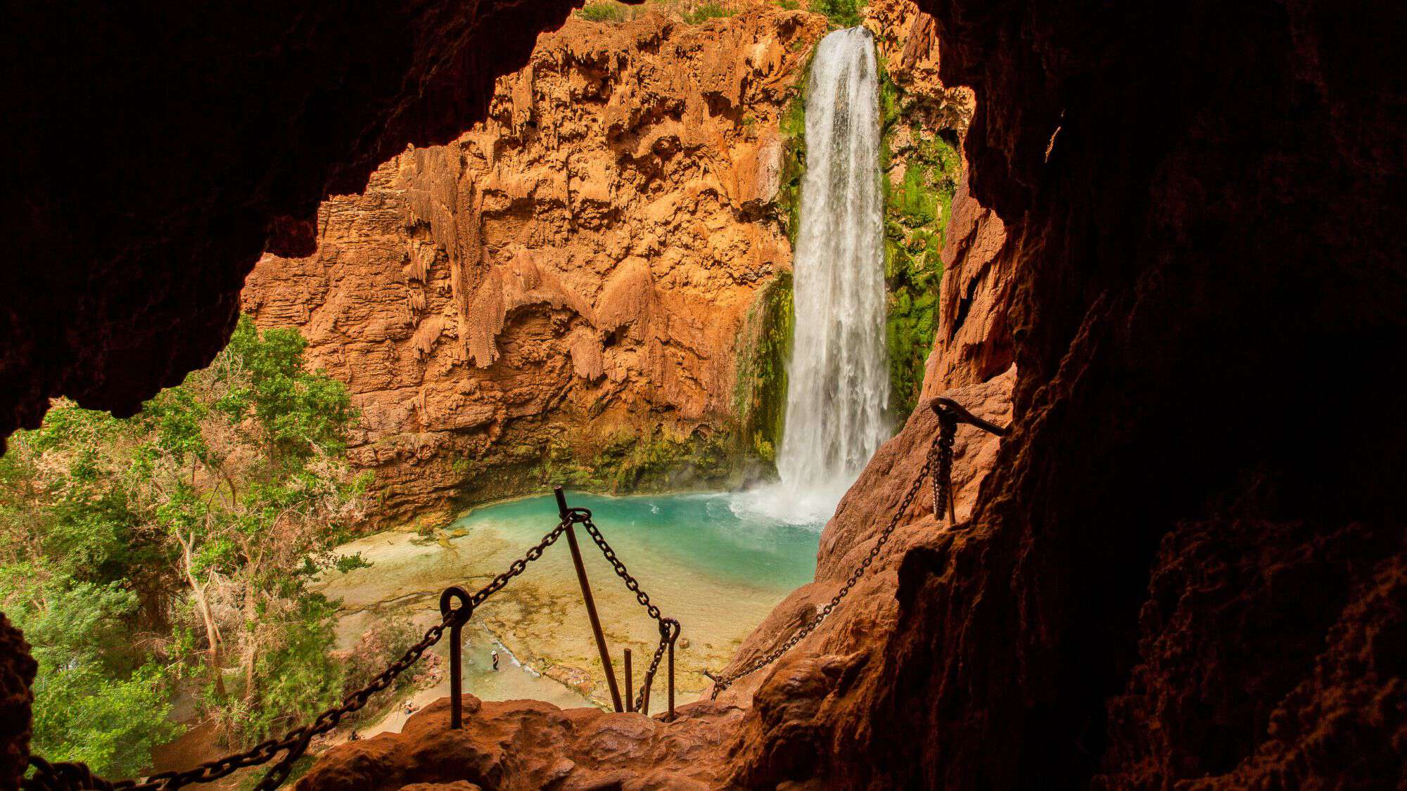 Looking out from a cave, this view captures Mooney Falls plunging into a turquoise pool below, with a chain-supported trail leading downward through the red rock.
