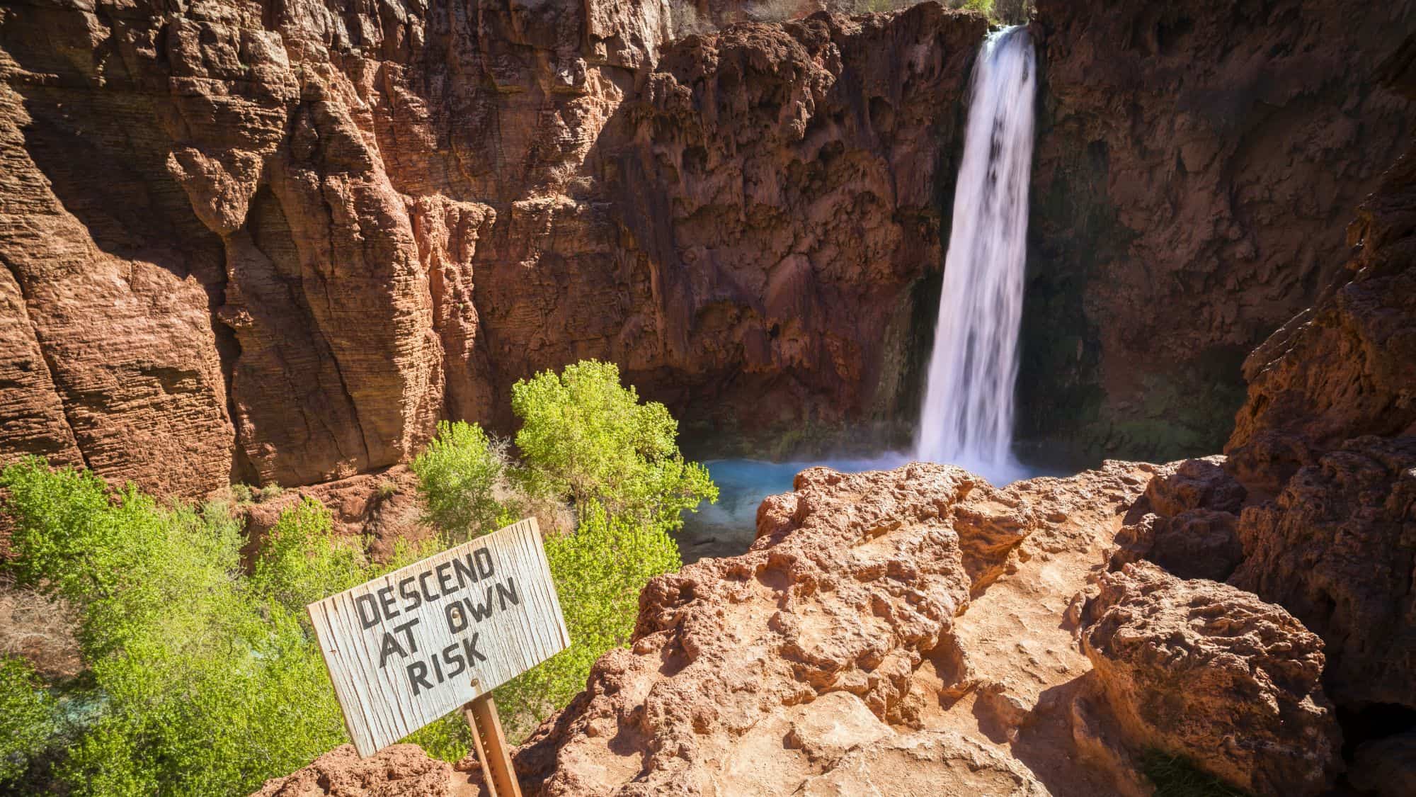 A dramatic view of Mooney Falls cascading into a blue-green pool at the base of towering red cliffs, with a weathered sign in the foreground reading “Descend at Own Risk.”
