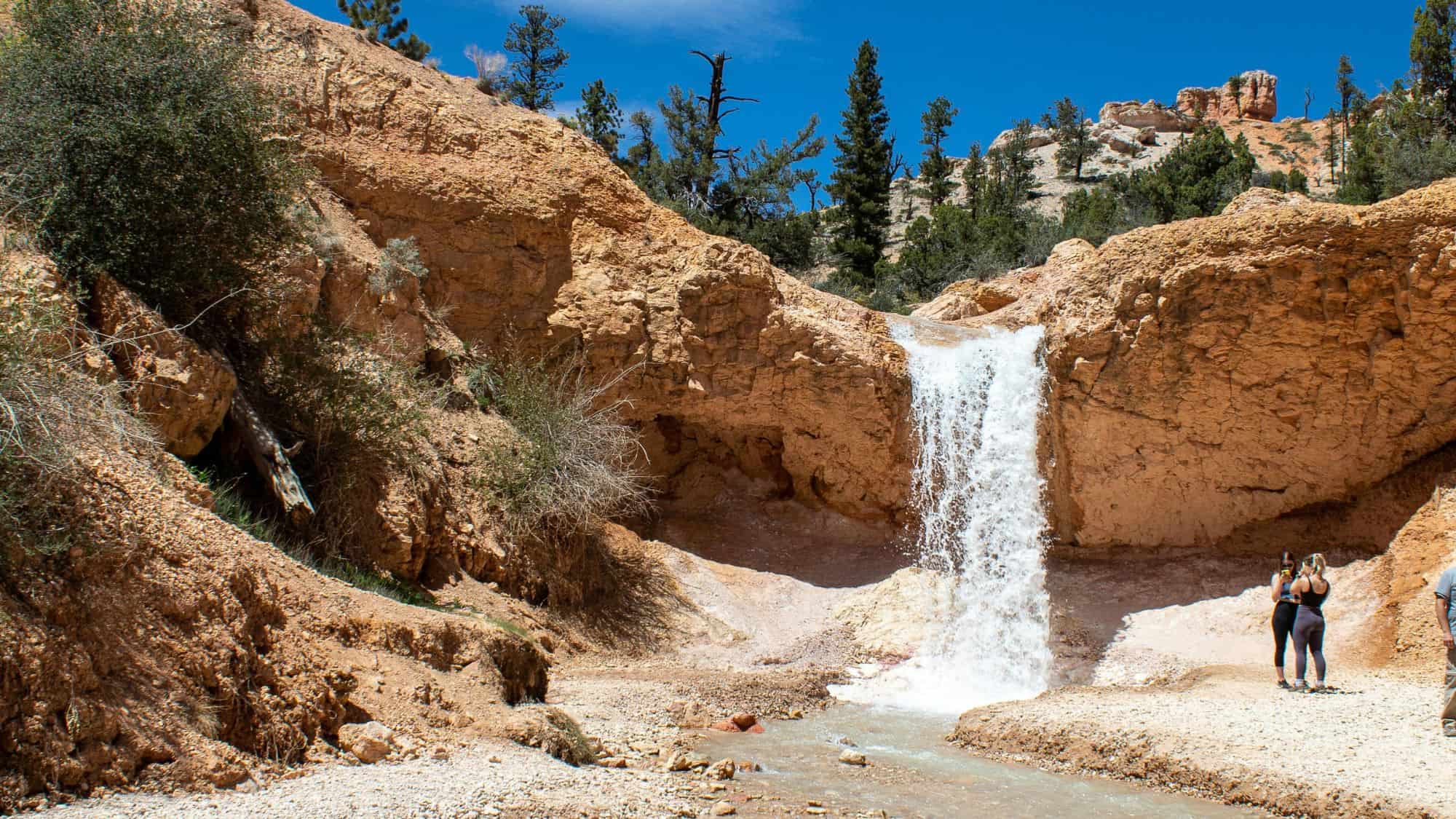 A small but picturesque waterfall cascades down a rocky cliffside in Bryce Canyon National Park. Two hikers stand nearby, admiring the desert oasis surrounded by red rock formations and sparse vegetation.