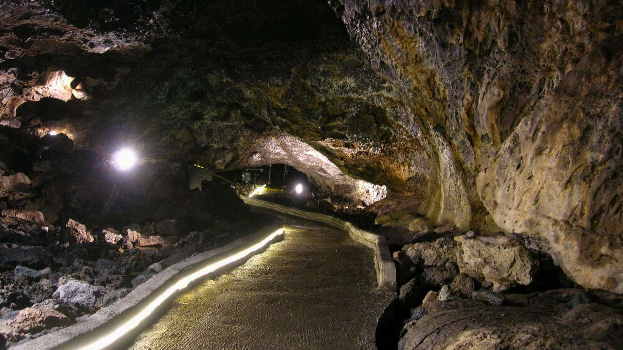 A dimly lit lava tube with a paved walkway illuminated by built-in lights. The rocky ceiling and walls display unique formations created by volcanic activity.