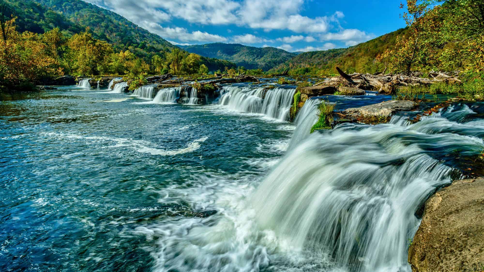 A series of cascading waterfalls flowing through a rocky river surrounded by lush green hills. The blue sky and rolling mountains create a serene, picturesque view.