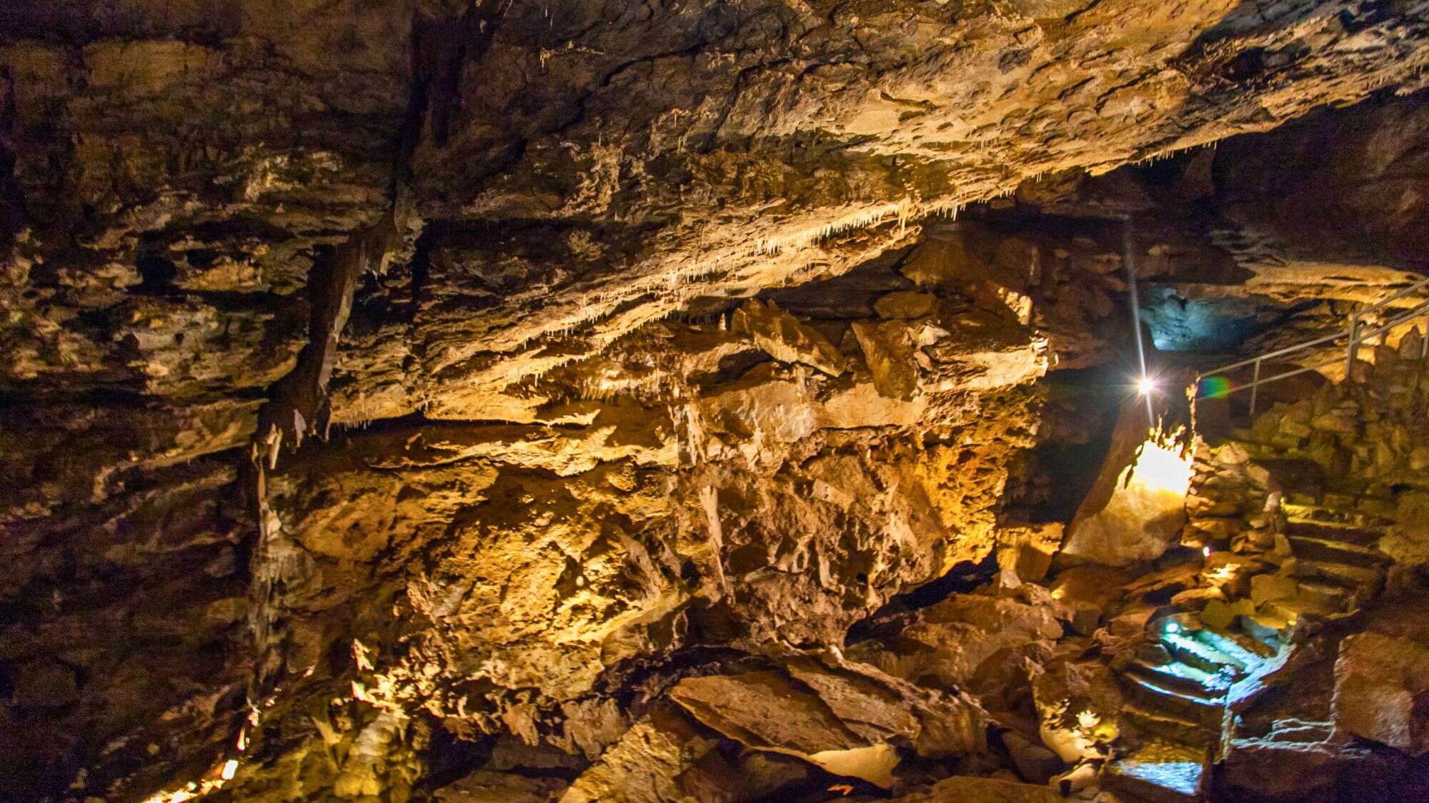 A cavernous passage with jagged rock formations and warm artificial lighting. A staircase leads deeper into the underground, revealing intricate stalactites and rugged cave walls.