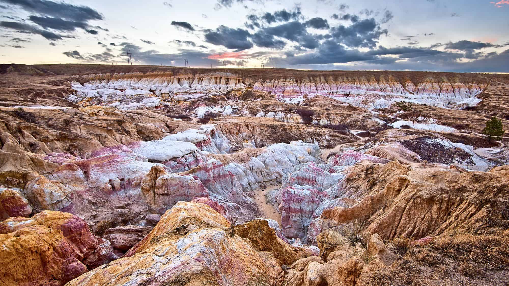 Otherworldly landscape of striped, pastel-colored rock formations with dramatic textures and hues, under a partly cloudy sky near sunset.