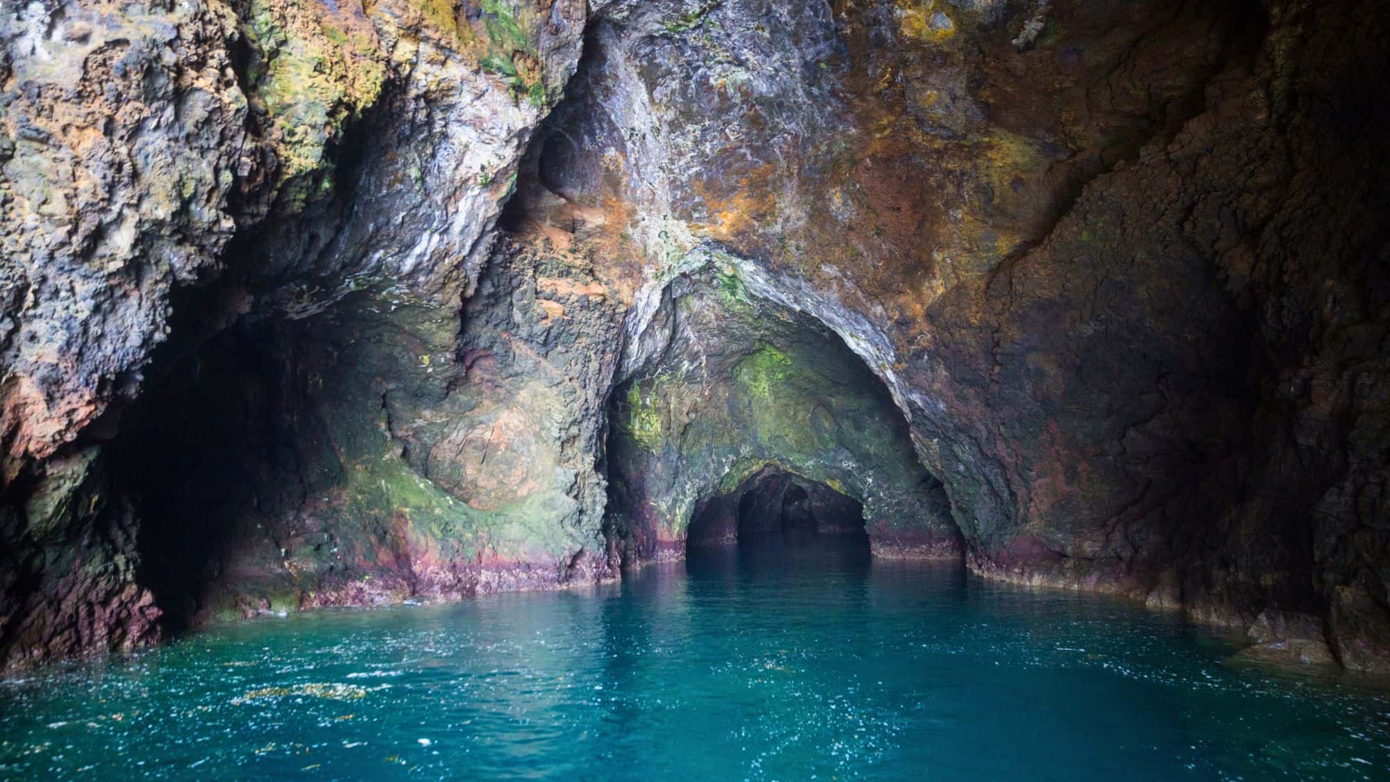 The entrance to Painted Cave on Santa Cruz Island reveals a vast, shadowy cavern with walls streaked in green, yellow, and rust-colored mineral deposits. The deep blue waters inside shimmer under soft natural light.