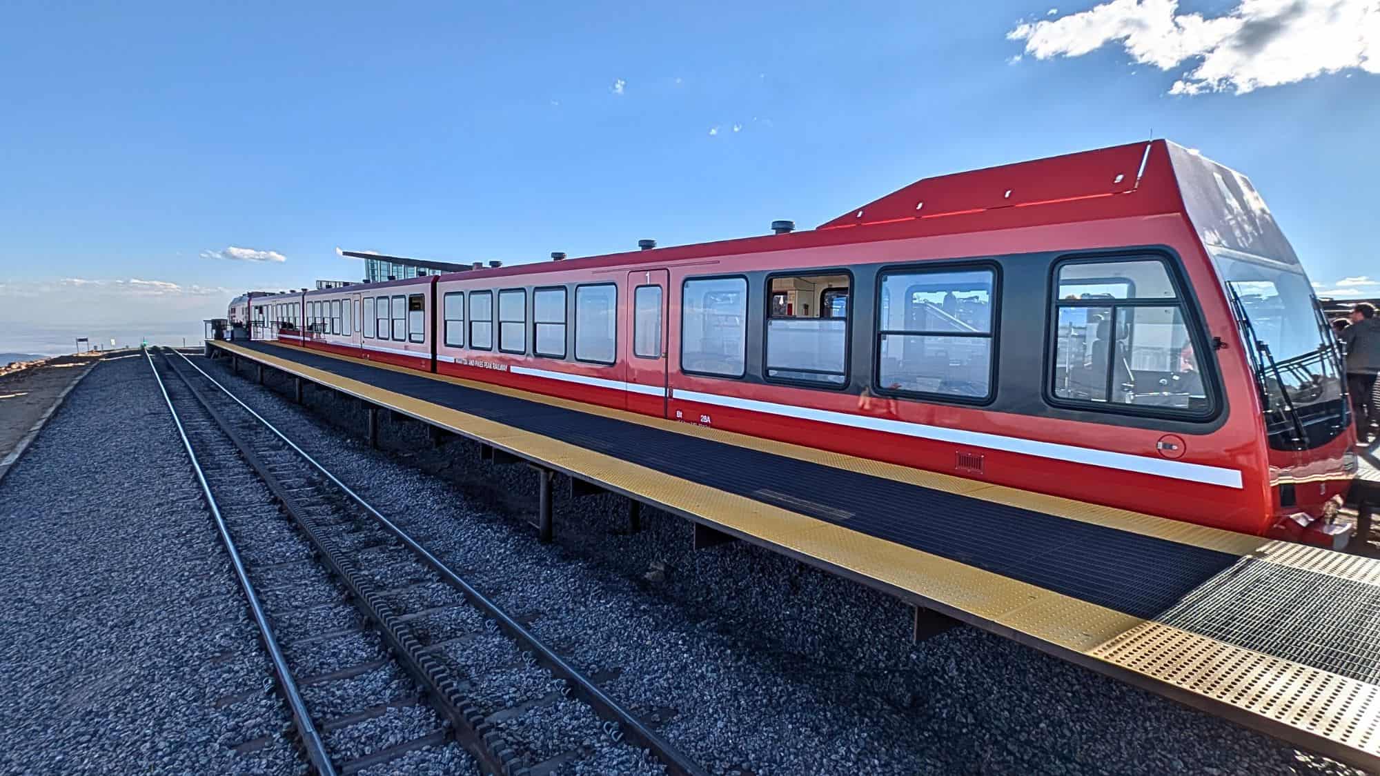 A red cog railway train sits at a mountain summit platform under a bright blue sky, with tracks stretching toward the edge of the peak.