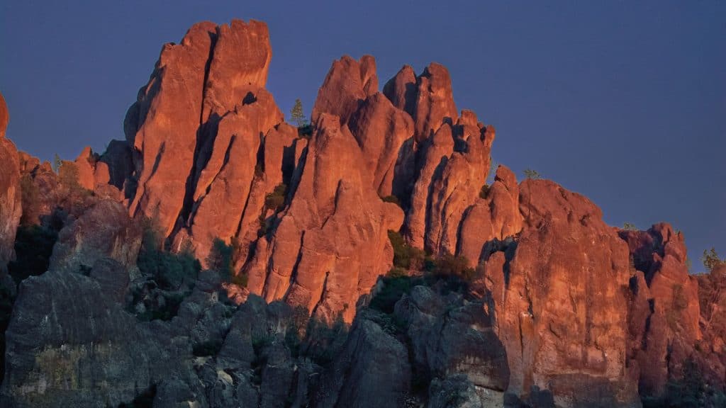 A dramatic view of the jagged rock formations at Pinnacles National Park, illuminated by the warm glow of the setting sun. The reddish-orange hues contrast against the deep blue sky, highlighting the rugged texture of the cliffs.