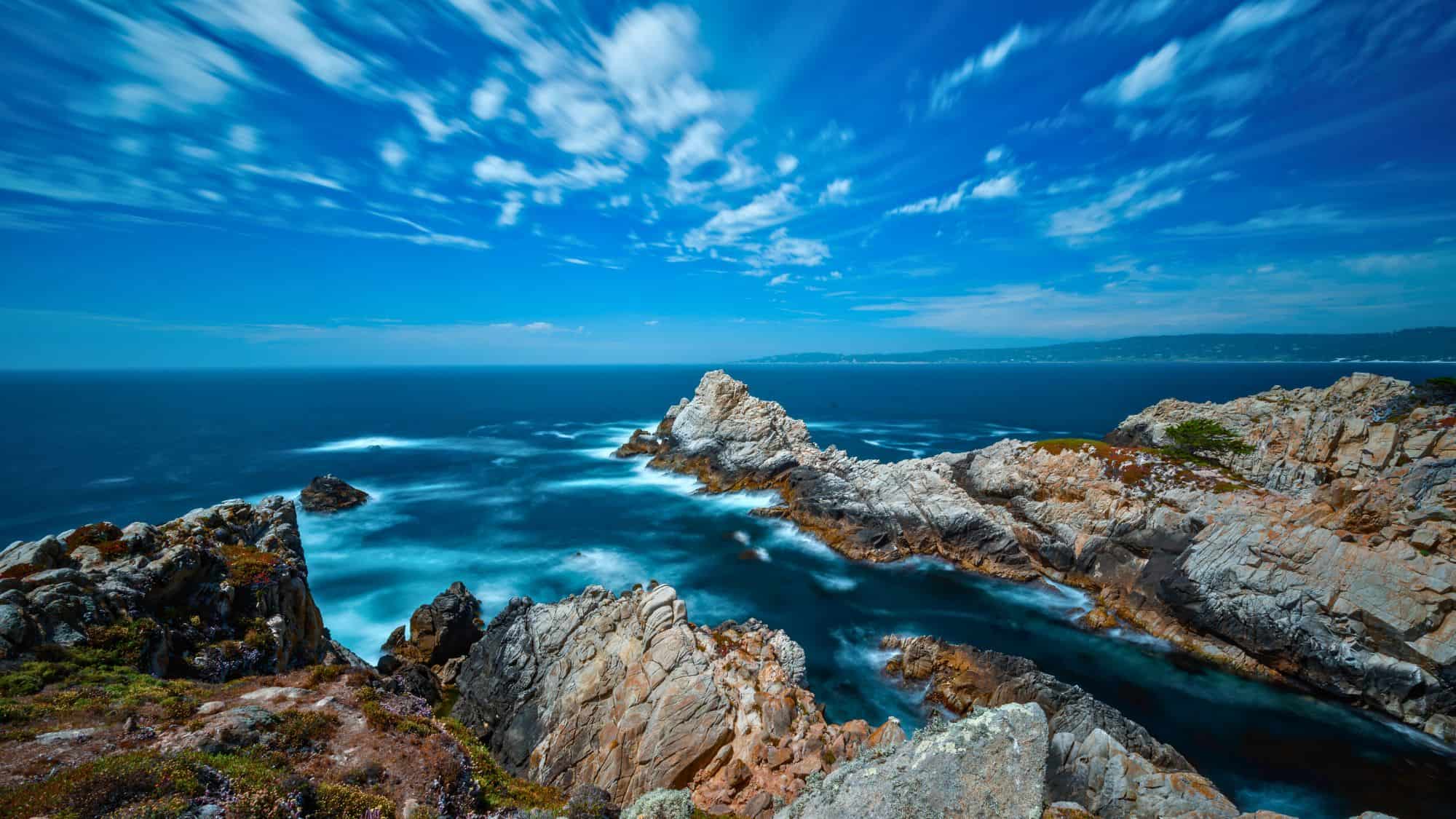 Jagged pale rock formations stretch into deep blue ocean waters, where long exposure waves swirl in soft white patterns around the shoreline. Above, streaked clouds sweep across a vivid sky, adding movement and drama to the expansive coastal view.