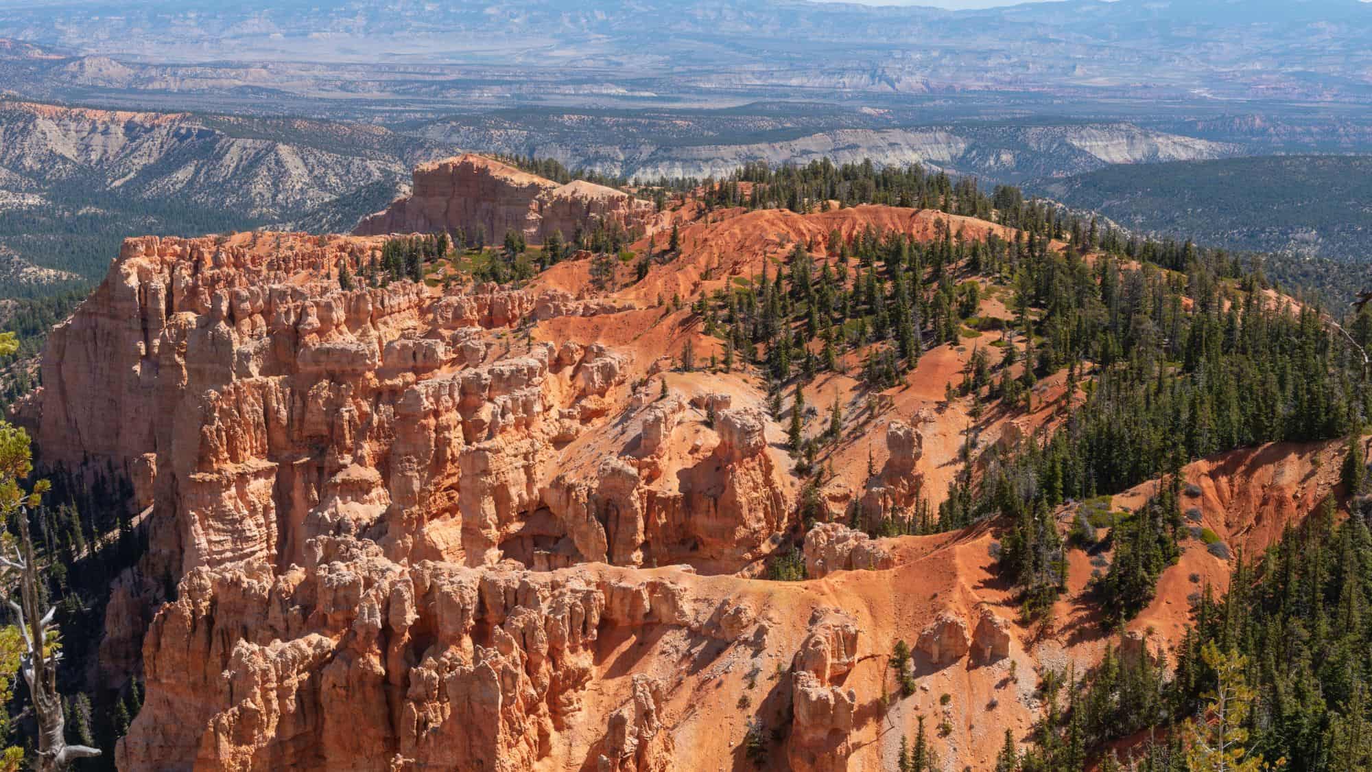 An aerial view of the vast Bryce Canyon landscape, showing dramatic cliffs, rolling plateaus, and scattered pine forests. The jagged red rock formations stand out against the blue sky.