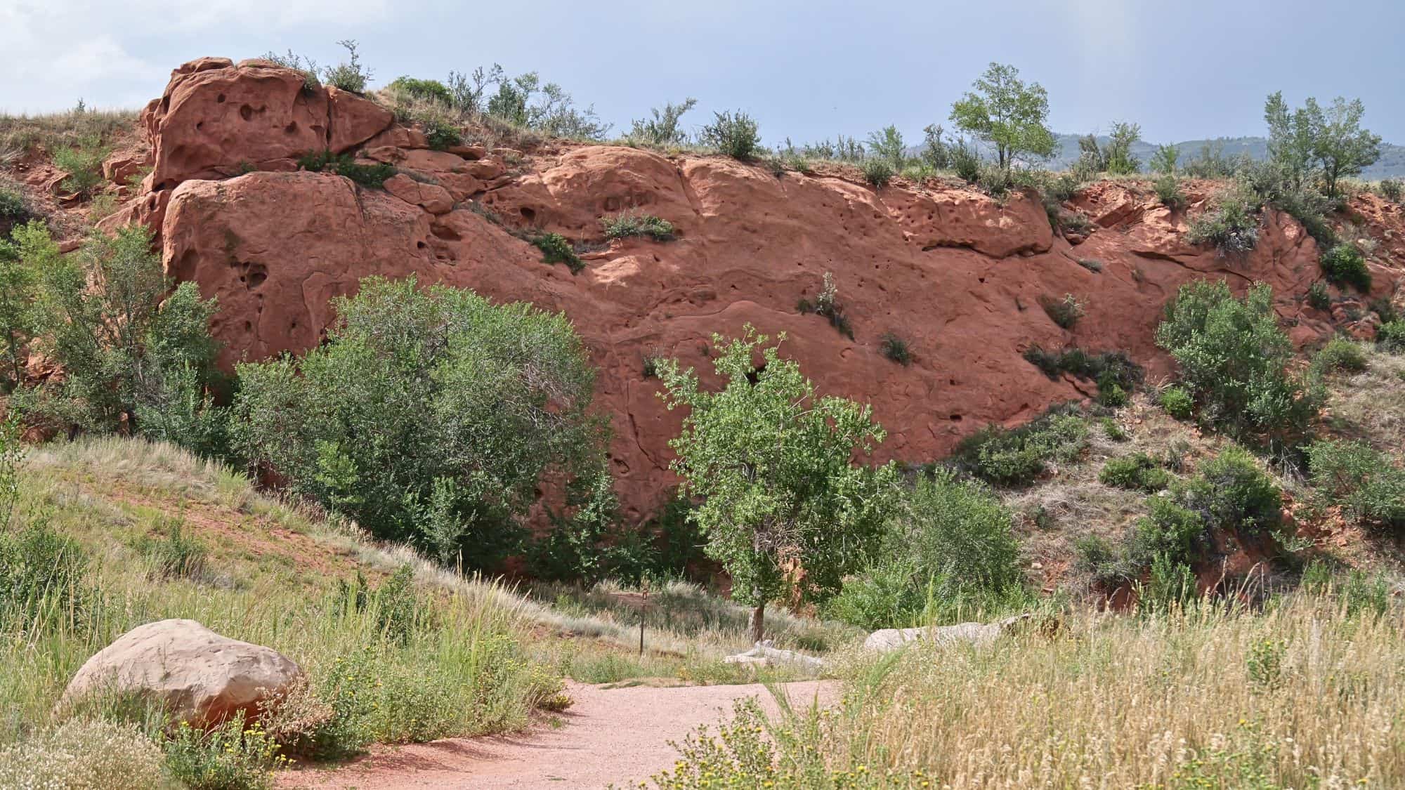 A reddish sandstone cliff with natural pitting rises above a grassy, tree-dotted trail in a scenic high desert environment.